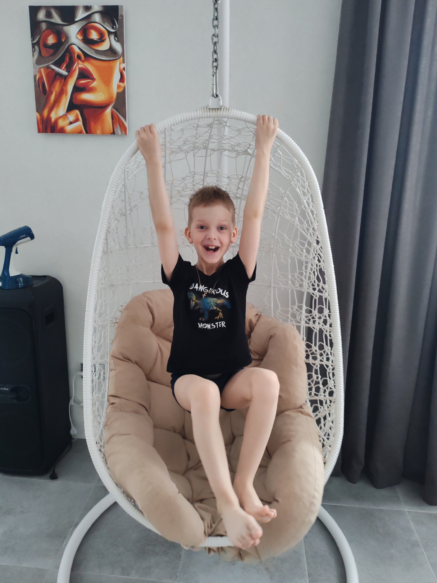A fair-skinned boy is seated in a white wicker hanging chair with a tan cushion. He is wearing a black t-shirt with the text "DANGEROUS MONSTER" on it, and black shorts. He has short brown hair and a necklace with a pendant. His arms are raised above his head and he is smiling with his mouth open.
To the left of the chair is a black speaker, and a blue clothes steamer is placed on top. On the wall behind the chair, there is a painting of a woman in a silver mask smoking a cigarette. To the right of the chair are gray curtains. The floor is tiled in gray. The location is Chaikovsky, Russia.