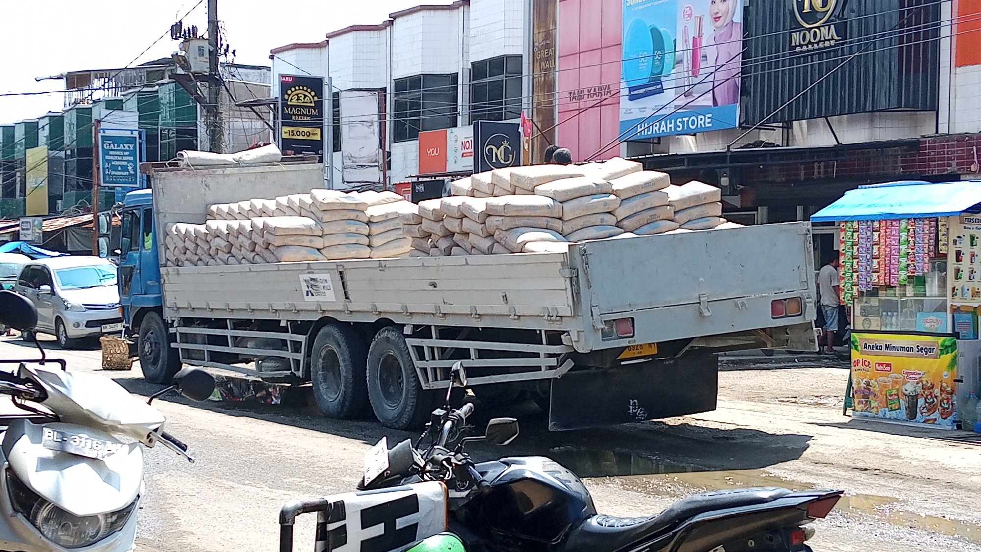 The image depicts a street scene in Lhokseumawe, Indonesia. A blue truck is loaded with numerous sacks of what appears to be cement or similar building material. The truck is parked on a street with a slight incline, and the ground appears wet in places, suggesting recent rain or a spill. In the foreground, a white motorcycle is partially visible. To the right of the truck, a small roadside stall with a blue awning is set up, selling various packaged goods and beverages. A man, wearing a grey t-shirt and shorts, stands behind the stall. In the background, several commercial buildings with storefronts and signage are visible. One prominent sign displays the text "HIJRAH STORE," and another reads "NOONA GALLERY." The overall atmosphere suggests a typical urban environment in Indonesia, likely a commercial or industrial area.