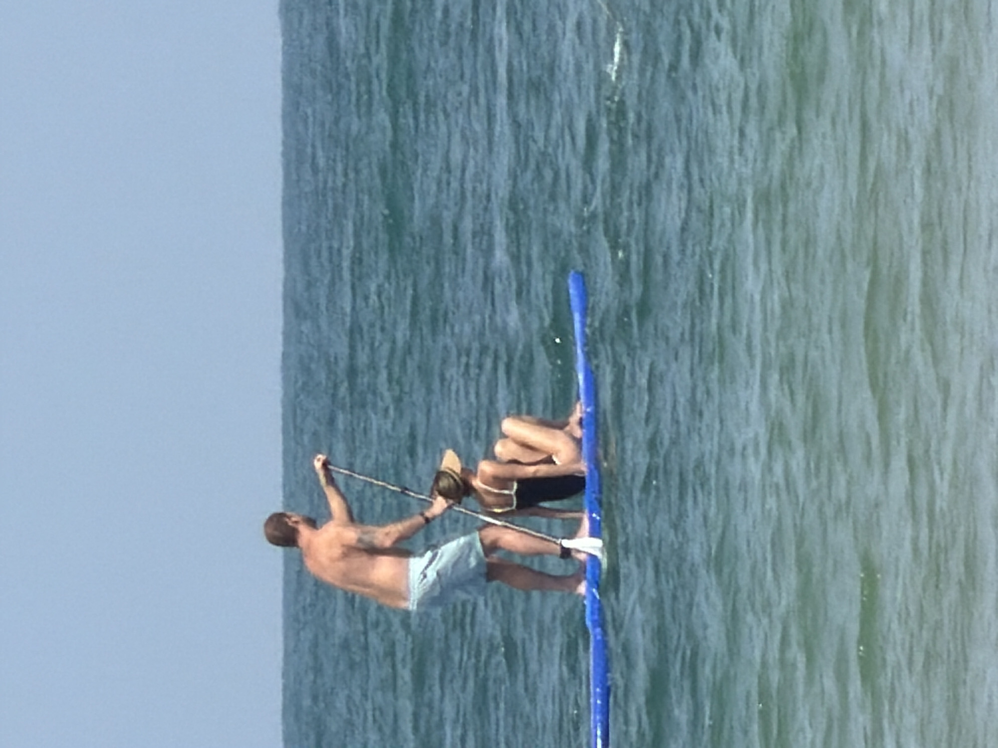 Two people are on a paddle board in the ocean off the coast of Tel-Aviv, Israel. One person is standing, shirtless, wearing light-colored shorts, and paddling. The other person is kneeling on the paddle board, wearing a bathing suit and sun hat. The ocean water is choppy. The sky is clear.