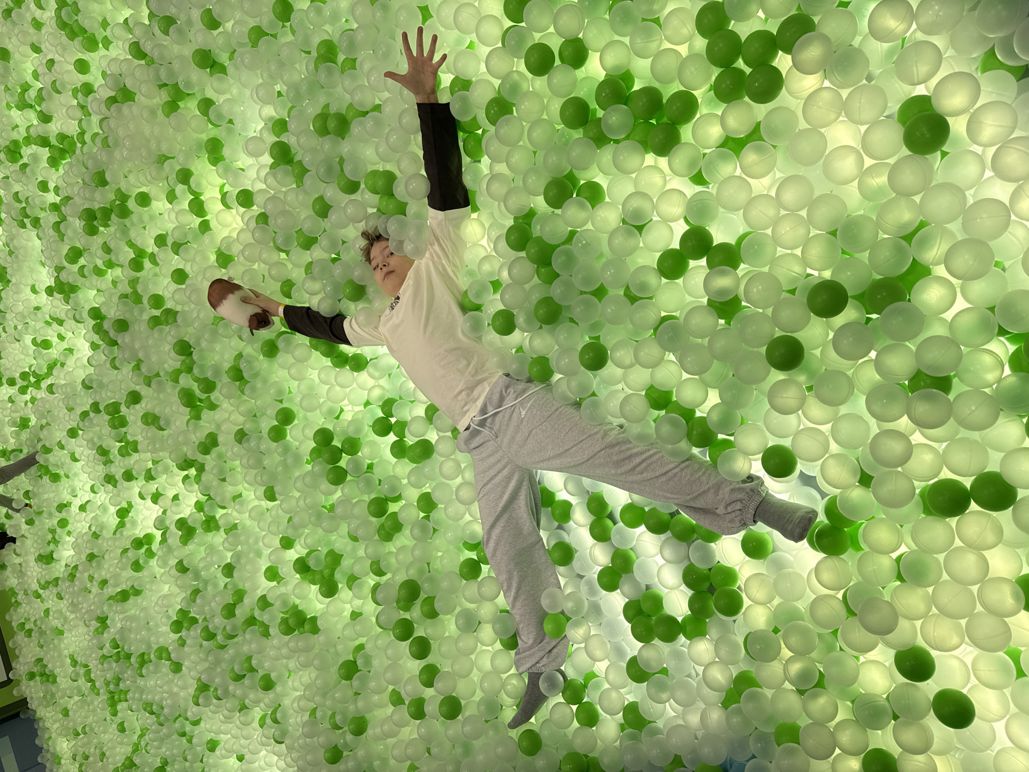 A young person lies on their back within a pit filled with green and white plastic balls. The person wears grey sweatpants, a white shirt with black sleeves, and grey socks. Their arms are extended; one hand holds a brown and white object, while the other hand is raised with fingers outstretched. The location is implied to be Перовское сельское поселение, Ukraine.