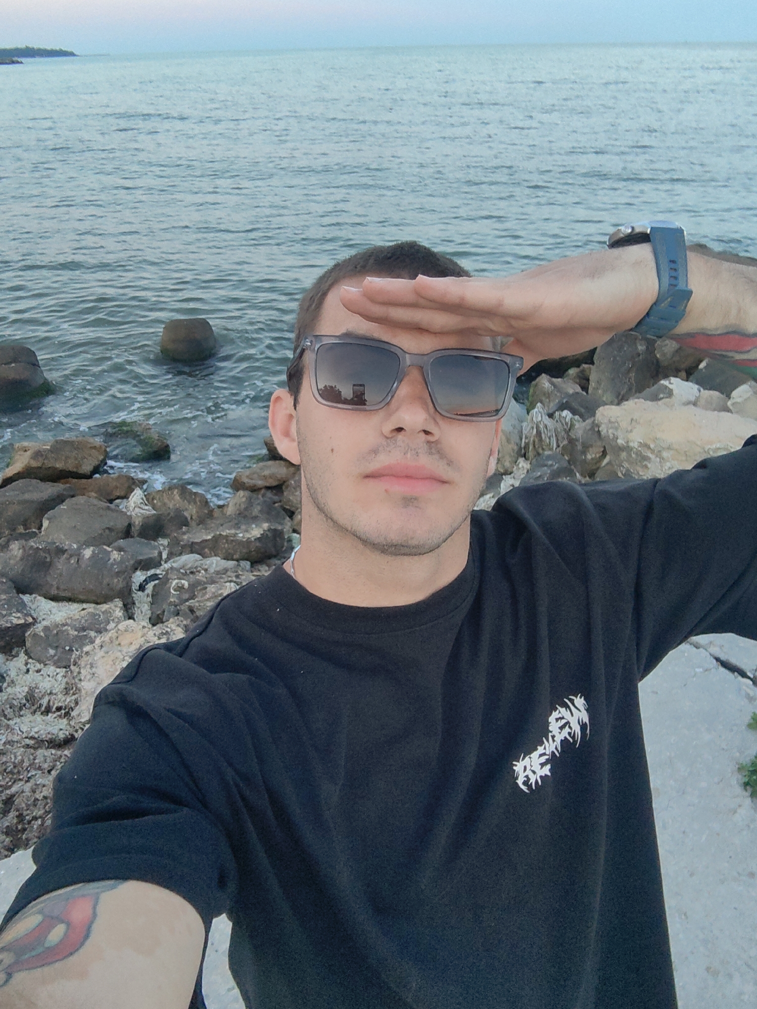 A young man with short dark hair, wearing sunglasses and a black t-shirt, is taking a selfie at the beach. He is standing on a rocky shore with the ocean in the background. The man is holding his hand up to his forehead, as if shielding his eyes from the sun. The water is calm and the sky is clear. The image is taken from a slightly low angle, looking up at the man. The t-shirt has a white graphic design on the left chest. He is wearing a dark blue digital watch on his left wrist. There is a tattoo visible on his left forearm. The overall setting appears to be a coastal area with a calm sea and a rocky embankment.