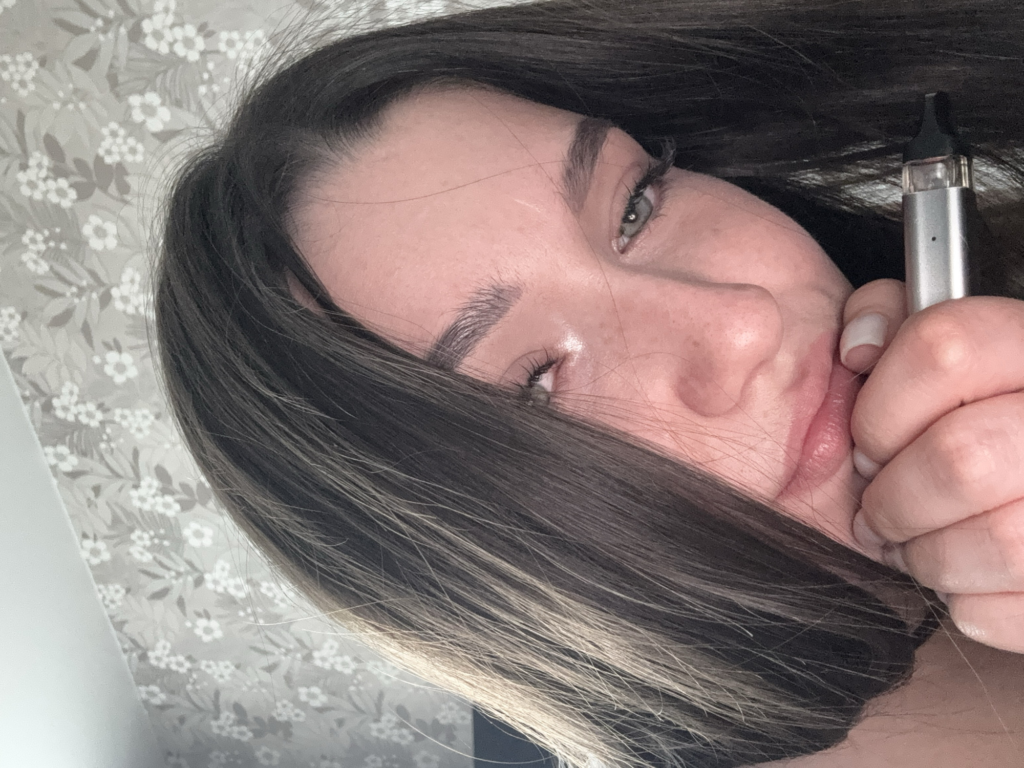 A woman with long, dark hair with blonde highlights, fair skin, and light-colored eyes is holding a silver vape pen in her right hand. The background is a patterned wallpaper with white floral designs on a gray background. The photo is taken indoors. No location information about the city of Perm, Russia is evident in the image.