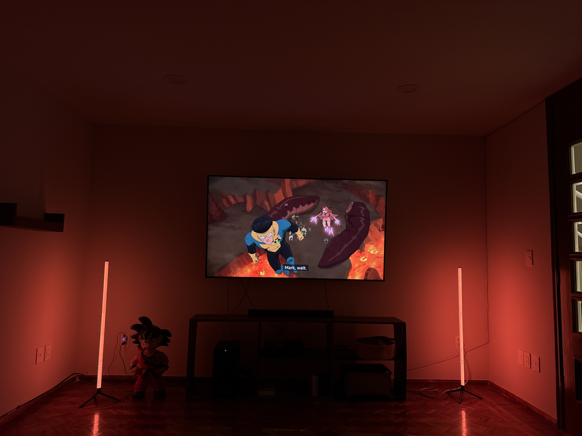 The image shows a dimly lit living room in Mexico City, Mexico, with a television displaying an animated scene. The room has a dark wooden floor and red-tinged walls illuminated by two floor lamps emitting a reddish light. The television is mounted above a dark entertainment console, which holds electronic devices and storage baskets. To the left of the console and near a floor lamp stands a Goku figurine. The lighting is dim.