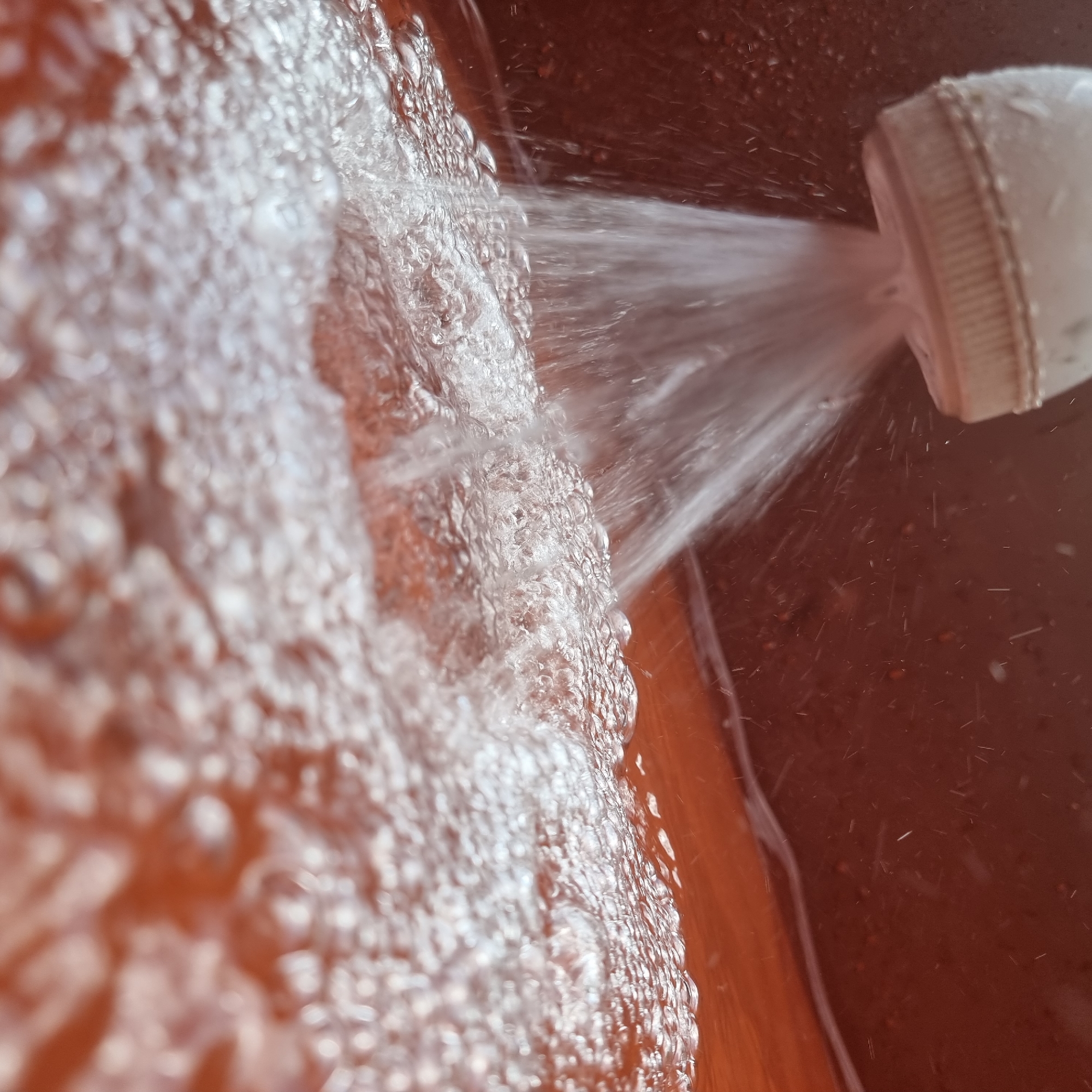 A close-up, low-angle shot captures a shower head releasing a stream of water into a body of water, likely a bathtub or sink. The shower head, white and textured, is positioned at the top left of the frame, with water spraying downwards. The water creates ripples and bubbles on the surface of the liquid. The background is a dark, reddish-brown color. The image is taken in Dunnae-myeon, South Korea.