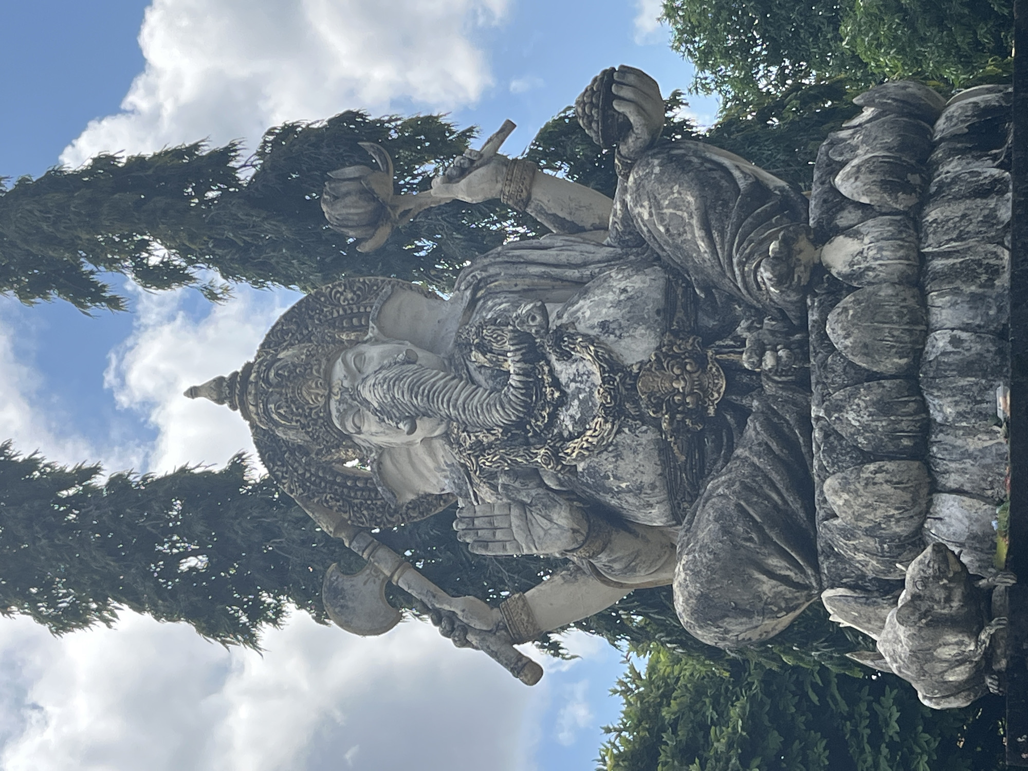 A weathered, stone statue of Ganesha, the elephant-headed Hindu deity, is depicted seated cross-legged on a lotus flower pedestal. The statue is ornate, with intricate carvings on its headdress, necklaces, and bracelets. Ganesha holds several objects in its multiple hands, including what appears to be a lotus flower and a bowl of fruit. To the left of the statue, a smaller stone carving of a mouse, Ganesha's vahana (mount), is visible. The statue is situated outdoors, with lush green trees and a cloudy blue sky forming the background. The overall impression is one of ancient spirituality and artistic craftsmanship. This statue is located in the city of Kuta Selatan, Indonesia, a popular tourist destination known for its temples and cultural heritage.