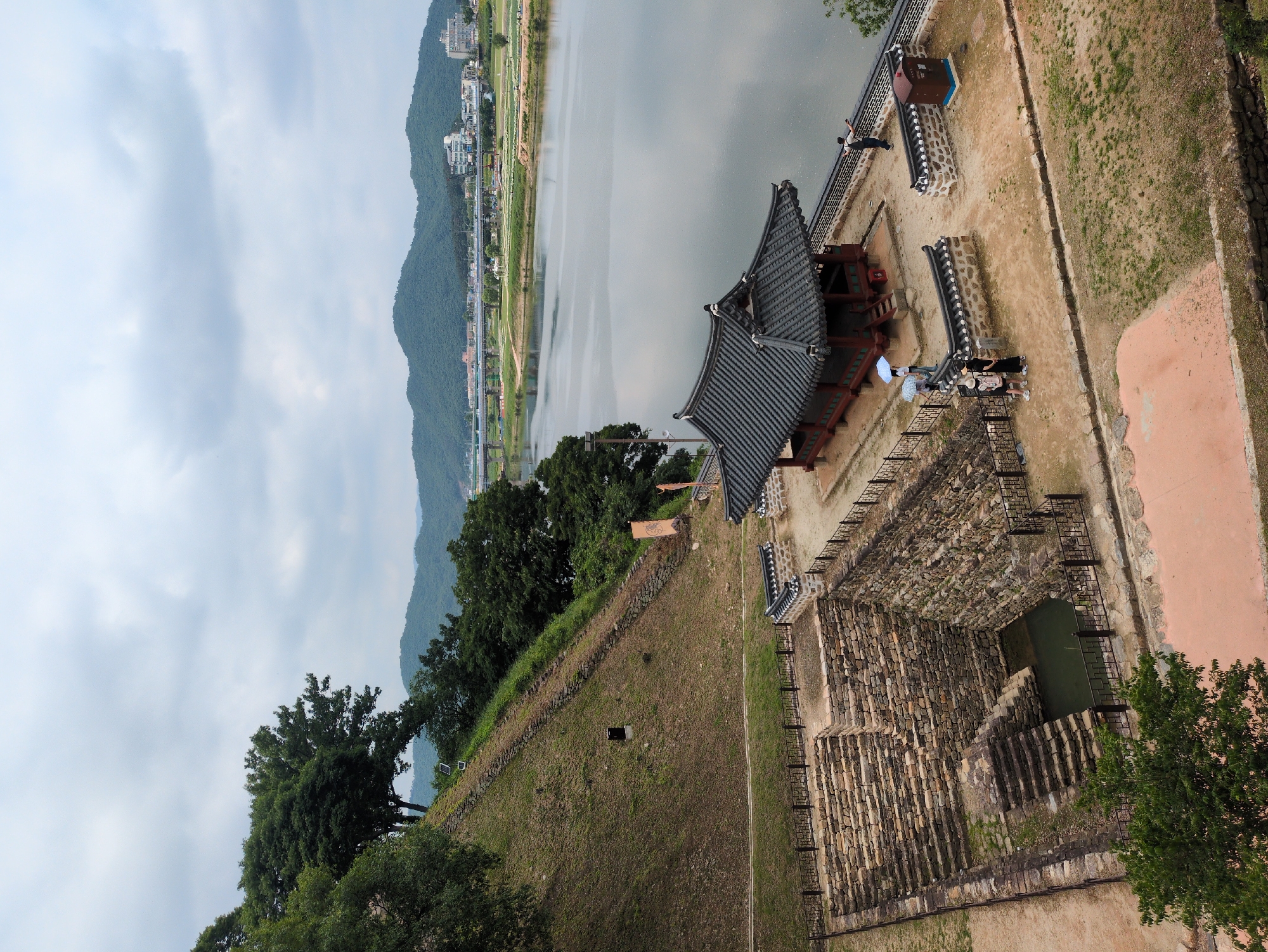 Here's a detailed description of the image:
The photograph, taken from an elevated perspective, captures a historical site with elements of architecture, landscape, and human presence. It depicts a scene likely located in Gongju-si, South Korea, suggested by the presence of traditional Korean architectural elements.
**Architectural Features:**
The image includes a building with a distinctive tiled roof, indicative of Korean historical architecture. Additionally, there's a stone-lined structure resembling a well or a reservoir. The landscape features stone fortifications and walls.
**Landscape:**
A body of water, potentially a river or lake, is visible in the background, flanked by mountains. The foreground includes grassy slopes, trees, and foliage, suggesting a natural setting integrated with the historical site.
**Human Presence:**
Several people are present on the grounds near the buildings. Some are standing near the stone fortifications, while others appear to be walking.
**Overall Environment:**
The scene is set under a cloudy sky. The lighting suggests it's daytime. The composition focuses on the interaction between architectural structures, natural surroundings, and human activity within a historical context.
The scene is an outdoor shot with a mix of natural and man-made elements within the context of a historical site in South Korea. The presence of people suggests the site is open to visitors.