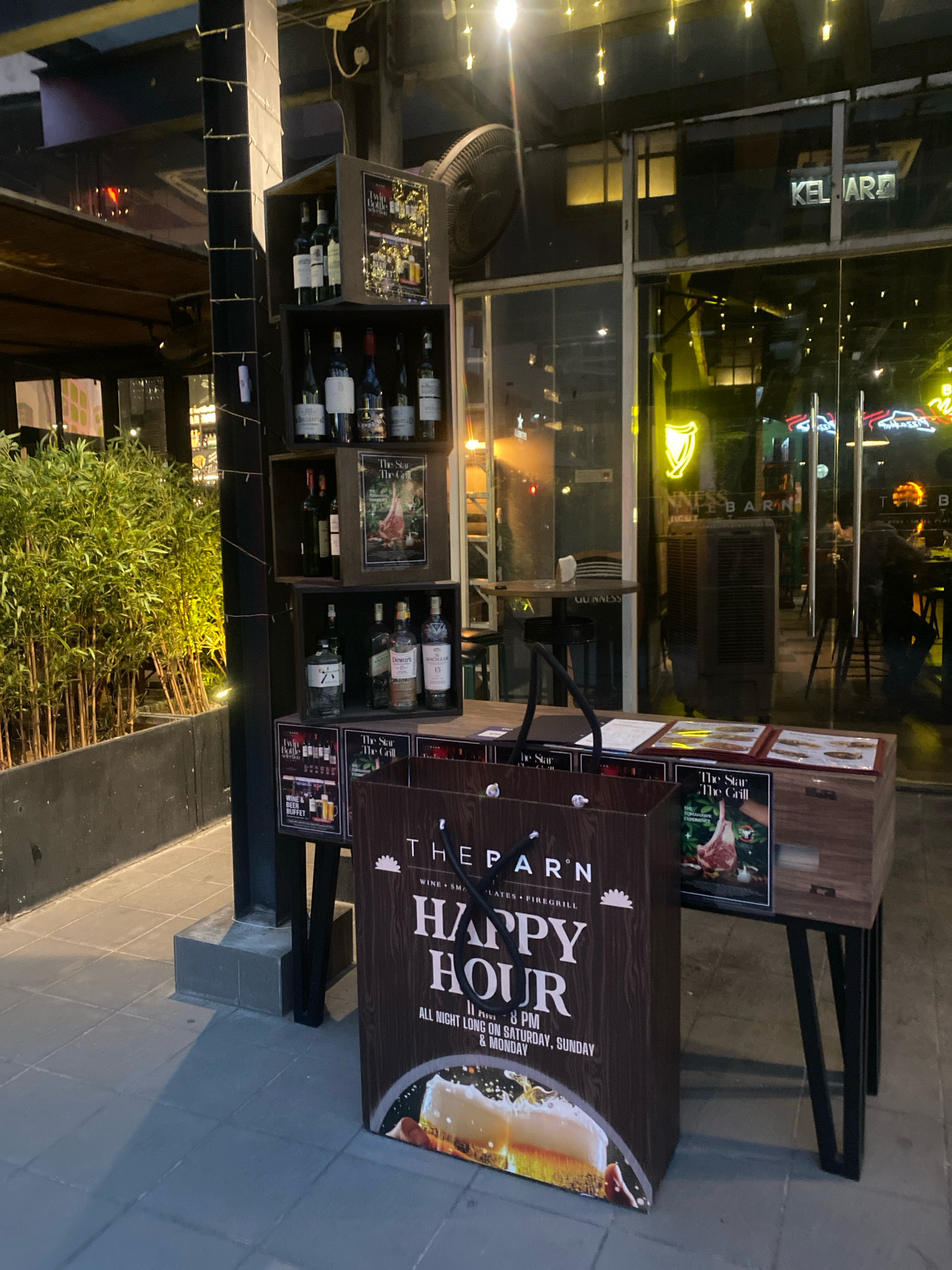 This image depicts the exterior of a bar or restaurant in Kuala Lumpur, Malaysia, at night. The establishment appears to be called "THE PARN" and has a "HAPPY HOUR" promotion advertised on a large, brown bag. Various bottles of alcohol, possibly wine and spirits, are displayed on shelves in wooden crates. One crate features a menu or advertisement for "The Star The Grill" with an image of a meat dish. Through the glass doors of the establishment, neon signs are visible, including a prominent Guinness logo. The scene includes outdoor seating with tables and chairs, as well as decorative lighting and lush green foliage in planters. The overall ambiance suggests a lively evening setting.