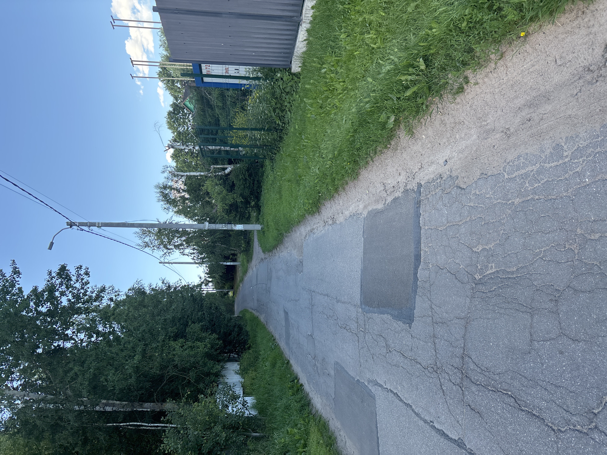 The image shows a moderately cracked asphalt road with several patches, suggesting it is in a state of disrepair. The road is lined with lush green grass and trees on both sides. On the right side of the road, there is a metal fence and a building with signage that appears to be in Russian. The sky is clear with a few white clouds. The scene appears to be a quiet street in a rural or suburban area. Based on the signage, the location is likely Yanino-2, Russia.