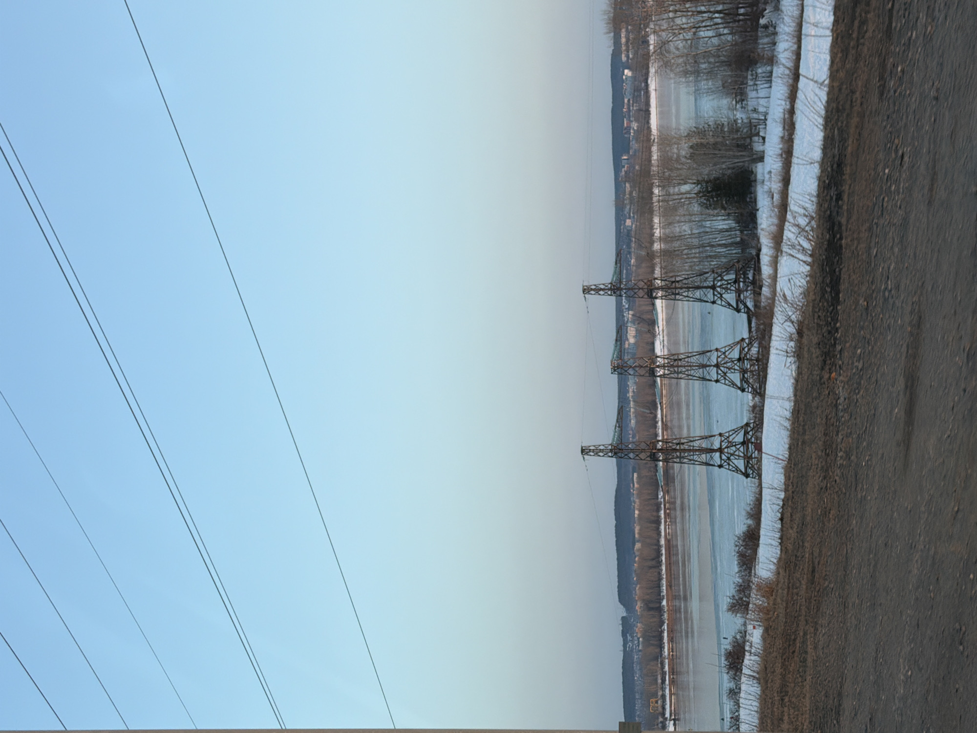 The image displays a wide landscape view in Noviy, Russia, under a clear, pale blue sky. The upper two-thirds of the frame consists of the sky, traversed diagonally by several thin, dark power lines. A light-colored vertical pole is visible along the extreme left edge, extending from bottom to top.
In the midground, a wide river or large body of water, partially covered by white ice and featuring areas of open water, spans horizontally. Three dark, lattice-structured metal transmission towers (pylons) are prominent, situated within or directly adjacent to the water. The immediate far bank of the river shows a sparse line of bare deciduous trees, some evergreen foliage, and scattered patches of snow.
Beyond the river and vegetation, a distant cityscape comprising various buildings is visible. This urban area is backed by a dark, forested hill range stretching across the horizon.
The foreground presents an uneven, dark brown surface, consistent with a dirt or gravel road or embankment. This area contains patches of dry, light brown vegetation and scattered snow closer to the river's edge. No human figures or discernible actions are present in the scene. The lighting indicates daytime.