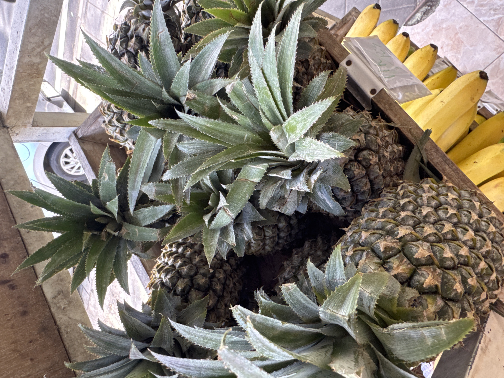 The image shows a display of pineapples and bananas in what appears to be a market setting, with the location inferred to be Kuta Selatan, Indonesia. The pineapples have spiky green leaves protruding from their tops. Adjacent to the pineapples are bunches of yellow bananas, some partially obscured by a pricing tag. The fruits are contained within a wooden frame, likely a display case or stall, and a vehicle wheel is visible in the background.