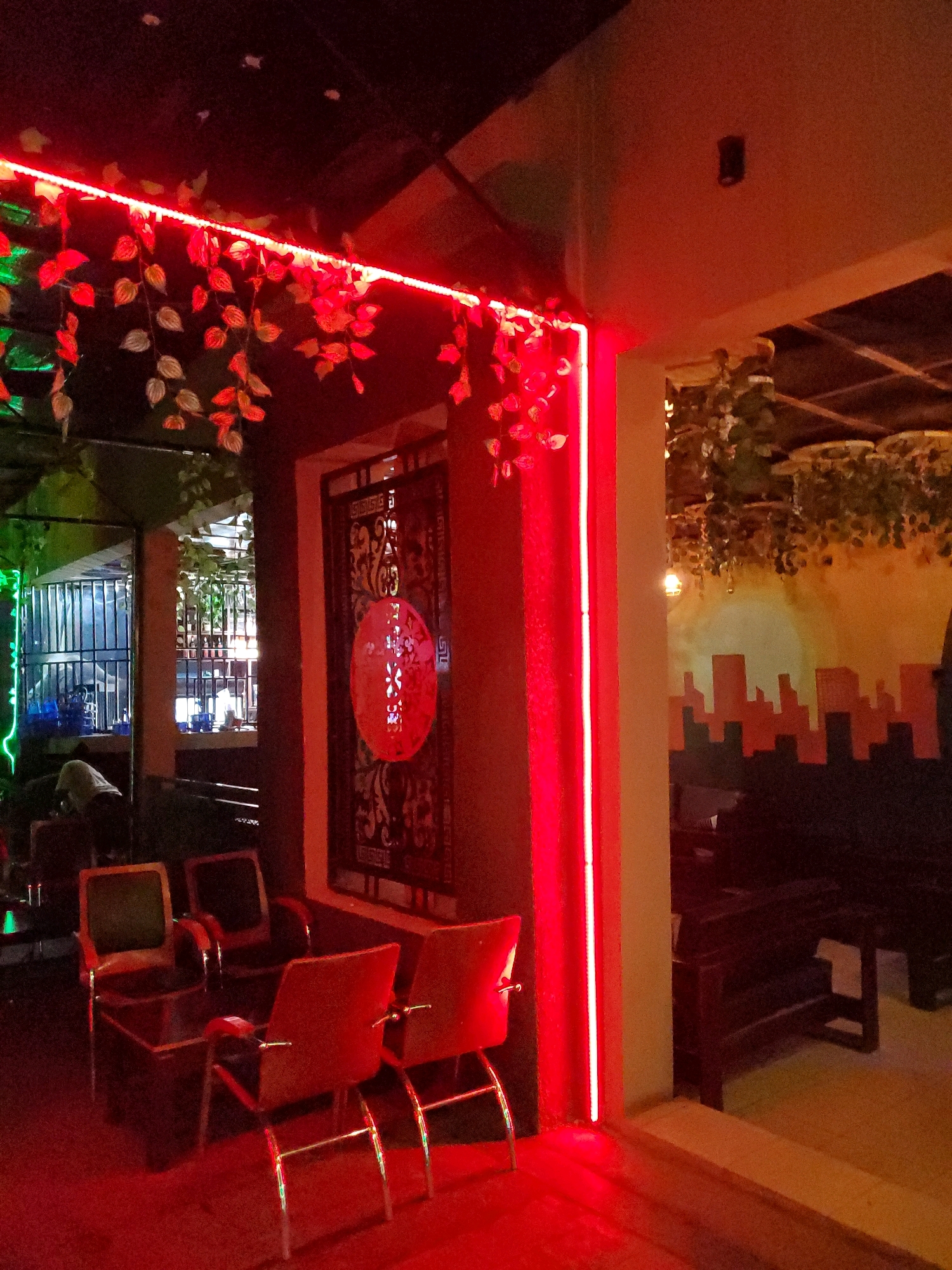 The image depicts the interior of what appears to be a lounge or bar area with a distinctly red ambiance due to the lighting. In the foreground, there are several chairs with red upholstery and metal frames, arranged around a dark wooden table. To the left, through an opening, a barred window is visible, with some greenery and lights. To the right of the chairs, there is a dark, ornate metal screen or door with a circular red design in the center. Further into the background, there are dark wooden benches and a wall with a mural of a cityscape under a yellow and orange sky. The overall impression is of a dimly lit, possibly entertainment-focused venue. The location is stated to be Jos, Nigeria.