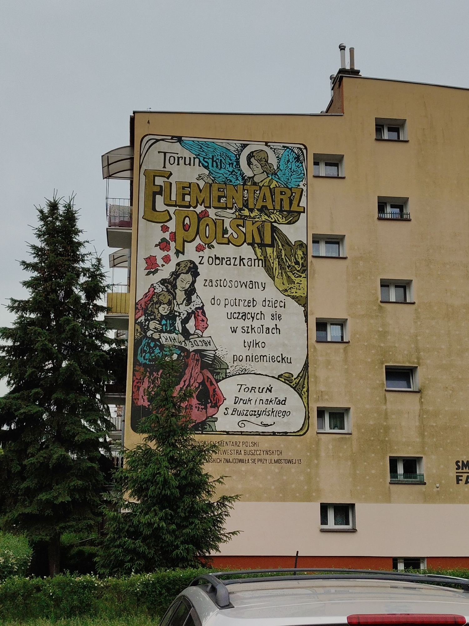 An external mural is painted on a beige apartment building. The mural is a large, colorful illustration. In the upper portion of the mural, the text "TORUŃSKI ELEMENTARZ POLSKI" is written in large, ornate, gold lettering. Below this, an illustration depicts a woman and a child reading a book. The woman is wearing a red and pink dress, and the child is wearing a blue and green dress. The book they are reading appears to have the letters "ABC" on it. Behind them, there are red roses and a depiction of an angel with blue wings. The mural is bordered by a thick, dark line. Below the main illustration, there is smaller text that reads "z obrazkami zastosowany do potrzeb dzieci uczących się w szkołach tylko po niemiecku" and "TORUŃ Druk i nakład S. Buszczyńskiego". Further below, there is text that reads "BUSZCZYŃSKIEGO ELEMENTARZ POLSKI KSIĘGARNIA WYDAWNICZA BUSZCZYŃSKA ZNAJDUJE SIĘ PRZY UL. MOSTOWEJ 13." A tall evergreen tree is positioned to the left of the mural, partially obscuring the building's facade and the mural itself. The sky is overcast and gray. The lower portion of the image shows the roof of a silver car and a glimpse of greenery. The building has several uniformly spaced windows.
