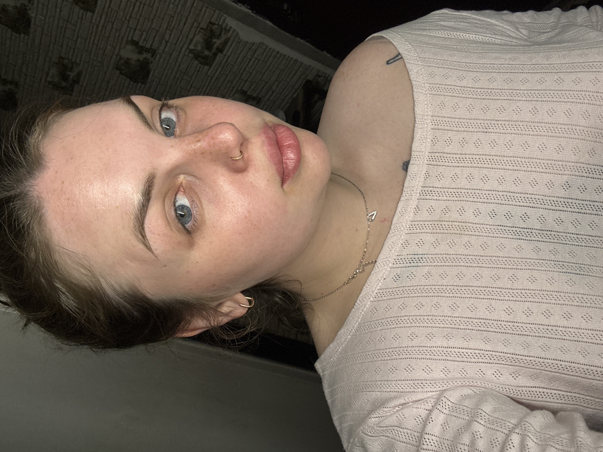 The image is a close-up portrait of a young woman, oriented with her head significantly tilted to the left. She has fair skin with visible freckles across her nose and cheeks, striking blue eyes, and dark brown hair that frames her face. Her full lips are tinted with a light pink color. She wears a small gold hoop nose ring and a matching small gold hoop earring on her visible right ear. A delicate gold chain necklace, featuring a small geometric pendant (possibly a 'V' or triangle), is visible around her neck.
She is dressed in a light-colored, likely off-white or very light pink, long-sleeved top. The top has a distinctive open-work knit pattern, characterized by alternating horizontal stripes of fine texture and rows of small diamond shapes. Visible beneath the sheer fabric on her right shoulder/upper arm are two tattoos: one appears to be a black paperclip or safety pin design, and another, darker, less defined mark below it.
The background is dark and indistinct, suggesting an indoor setting with a subtle, dark patterned wall. The lighting appears artificial, implying the photo was taken indoors, possibly at night. Her expression is neutral and she looks directly at the viewer. No text is visible in the image. The location context is Chornomorskoe, Ukraine.