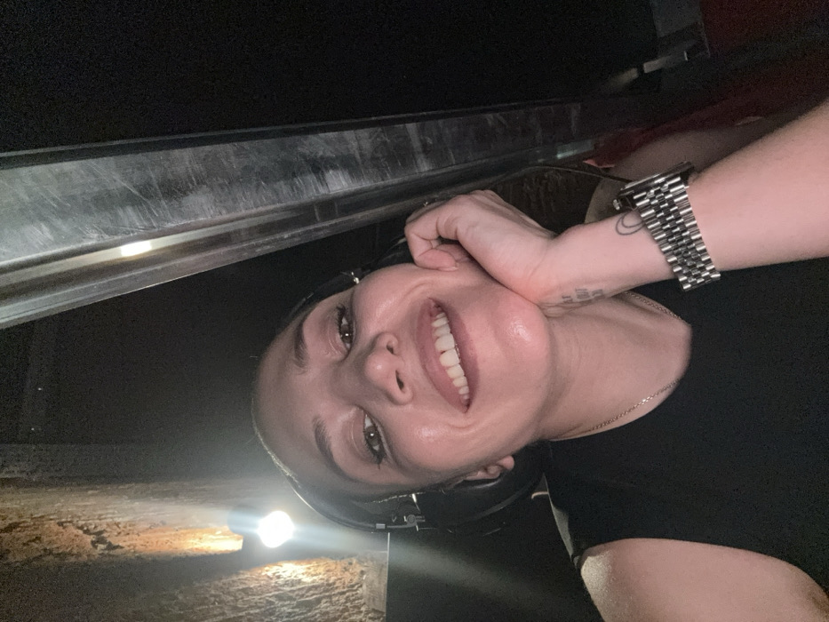 A woman is depicted in a close-up, upward-angled selfie, smiling broadly and looking directly into the camera. She appears to be an adult, possibly in her 30s-40s, with dark hair, dark eye makeup, and a fair complexion. She is wearing black over-ear headphones, a dark sleeveless top, and a thin chain necklace. Her left hand supports her chin/jawline, and a metallic, link-style watch is visible on her left wrist. Textual ink is present on her left inner forearm. The background is dimly lit and suggests an indoor location, likely an entertainment or production venue in Moscow, Russia. To the left, a textured, rough-surfaced wall, possibly concrete or stone, is illuminated by a single exposed light bulb. To the right, a vertical, scratched, metallic or dark reflective panel or beam is present. No specific landmarks or unique identifiers are visible to pinpoint an exact address or named venue within Moscow. The overall environment suggests an industrial or backstage setting within such a venue.