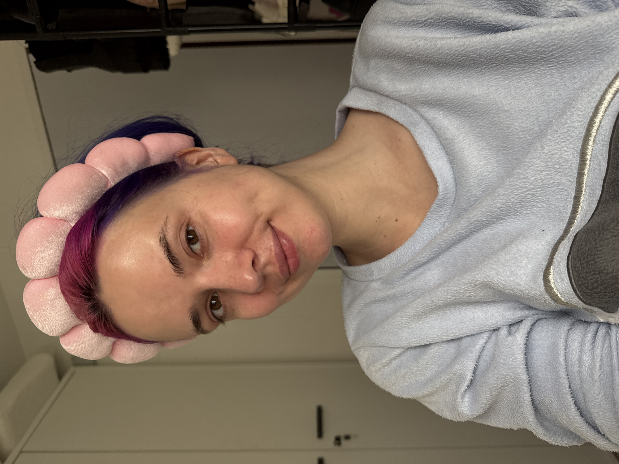 A person is seen indoors, likely in a domestic environment, wearing a light blue fleece top and a pink flower-shaped headband. The person has dyed hair that transitions from purple/magenta at the front to blue towards the back. The background features a plain white wall with what seems to be a built-in cabinet or wardrobe on the left side. Additionally, there is a darker shelf above the person in the background. The location is likely in the City of Zagreb, Croatia.