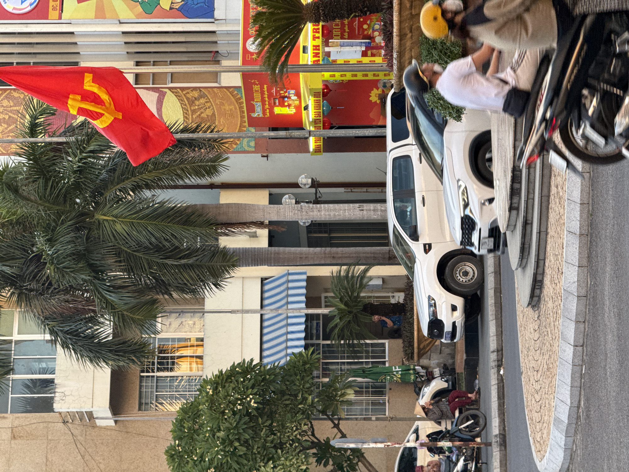 The image depicts a street scene in Nha Trang, Vietnam, captured during the daytime. In the foreground, a circular paved island with granite edging separates roadways. Two motorcycles are in motion on the right side of the frame. The rider of the foremost motorcycle wears a black helmet and a white t-shirt, while the passenger of the second motorcycle is partially visible, wearing a tan jacket and a yellow helmet.
Behind the island, a white pickup truck is parked on the left, facing left. To its right, a white SUV is parked, facing right. On the sidewalk to the left of the truck, a person wearing a conical hat and dark clothing sits on a motorcycle, drinking from a cup.
The background features a building with several windows. A red flag with a yellow hammer and sickle emblem, characteristic of Vietnam's ruling party, flies from a flagpole in front of the building. Palm trees are prominent in the scene, with one tall palm tree obscuring part of the flag. A blue and white striped awning extends from the building above a storefront. Signs are visible on the buildings, some with Vietnamese text and colorful graphics, indicating businesses. One sign displays "Sanest MOON CAKE". The overall environment suggests an urban commercial area.