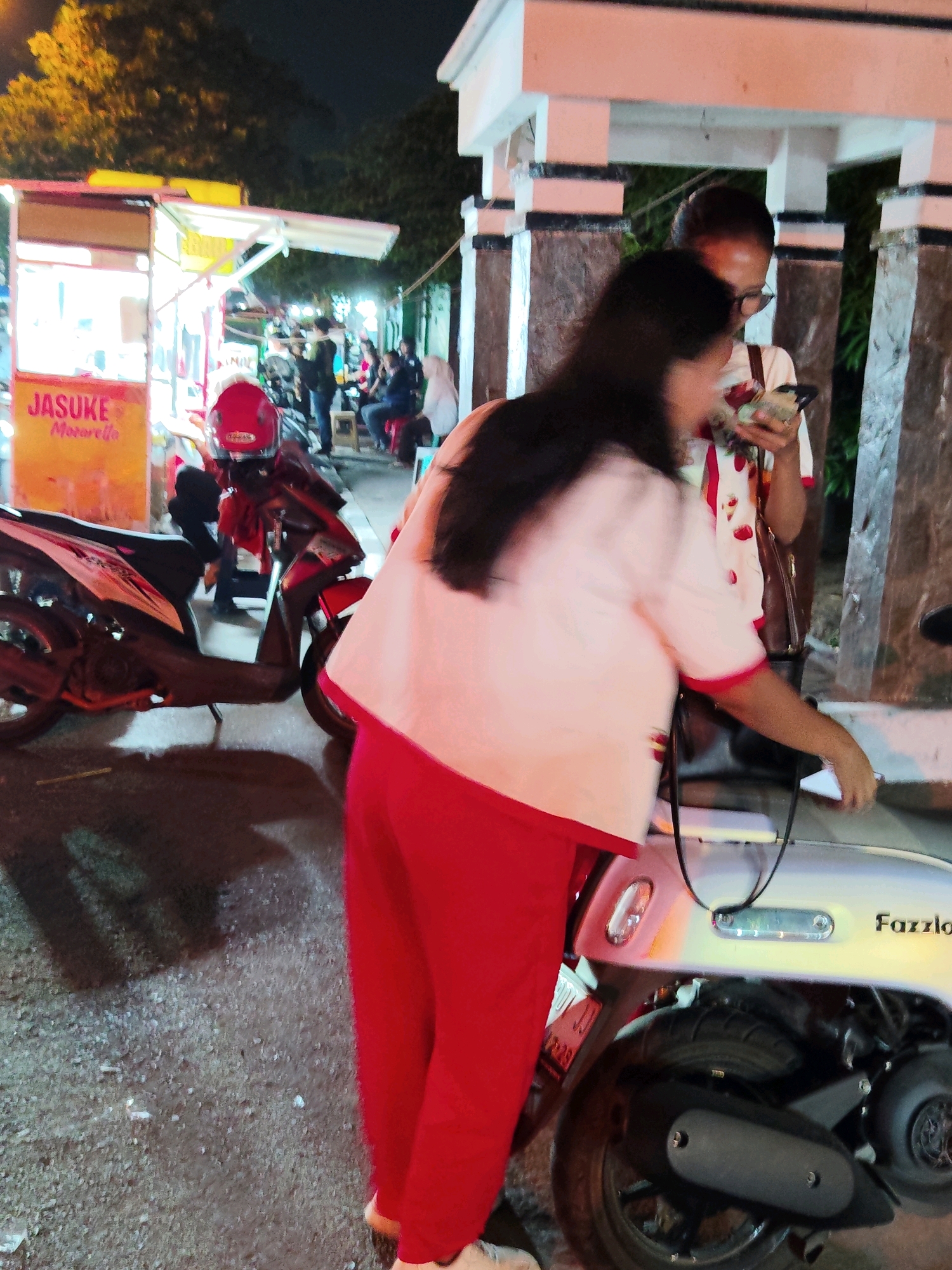In a street scene in Purwakarta, Indonesia, at night, a person wearing a pink top and red pants is interacting with a light-colored scooter. To the left of the scooter, another scooter with a red helmet on its seat is parked. Behind them, a food stall with "JASUKE" and "Motorella" written on it is visible. In the background, several people are gathered, some sitting on chairs. The environment suggests a bustling night market or street food area with artificial lighting illuminating the scene.