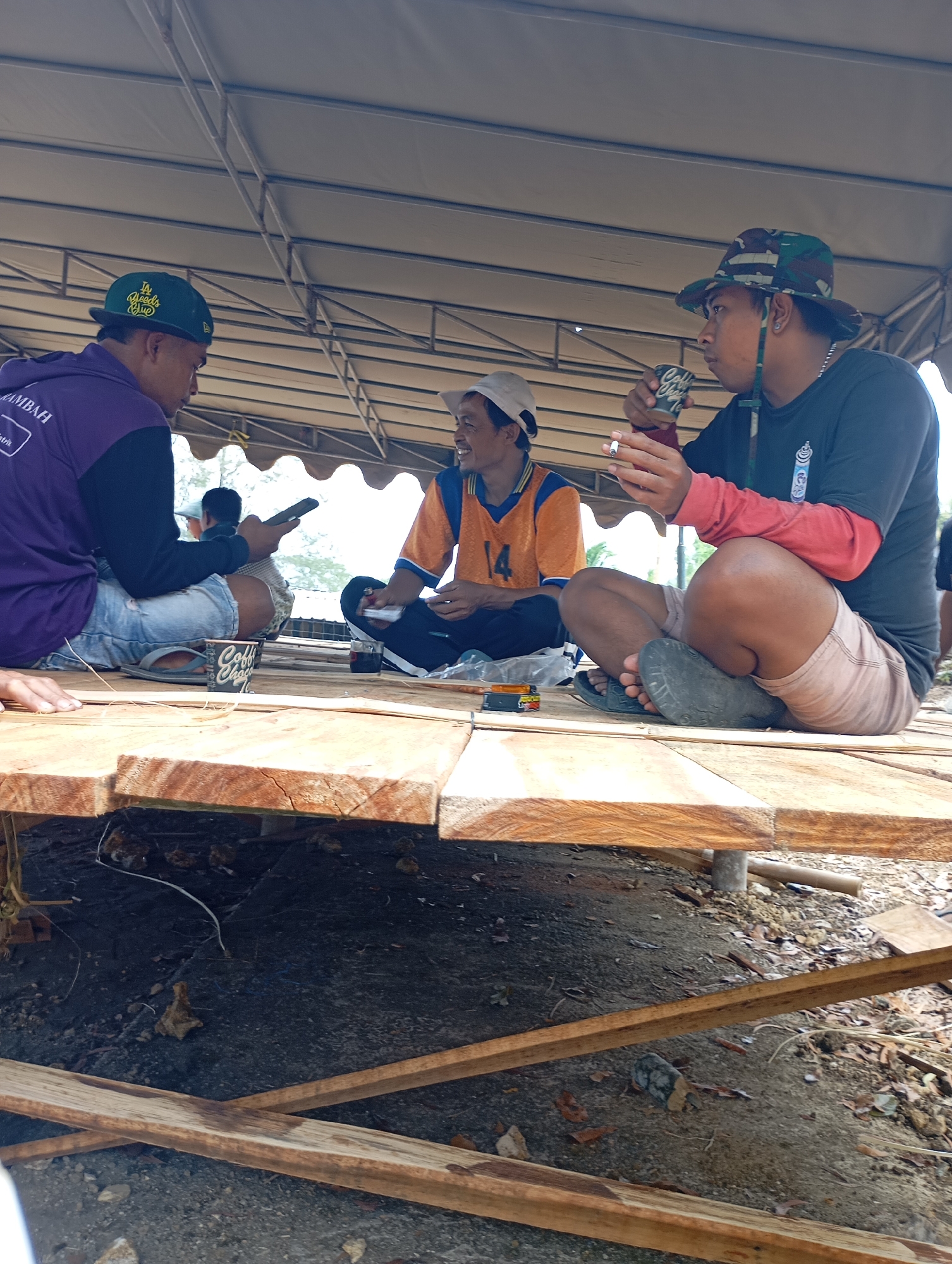The image depicts three men sitting on a wooden platform outdoors, likely in Ketepung, Indonesia. The scene is under a large, tan-colored tent or canopy, supported by metal beams.
In the foreground, a man wearing a purple hoodie and blue jeans is looking at his phone. A coffee cup sits on the platform near him. To his right, a man wearing an orange and blue shirt with the number "14" is holding a cigarette and looking down. Next to him, a man in a black t-shirt with a red sleeve and a camouflage hat is holding a cigarette and a small cup, appearing to be drinking from it. His legs are crossed, and he is wearing light-colored shorts.
The wooden platform appears to be under construction or recently built, with rough-hewn planks. The ground beneath the platform is composed of dirt and scattered debris, including leaves and small rocks. In the background, beyond the tent, glimpses of foliage and a body of water are visible, suggesting a rural or semi-rural outdoor setting. The overall atmosphere seems casual and relaxed, with the men engaged in conversation or individual activities.
