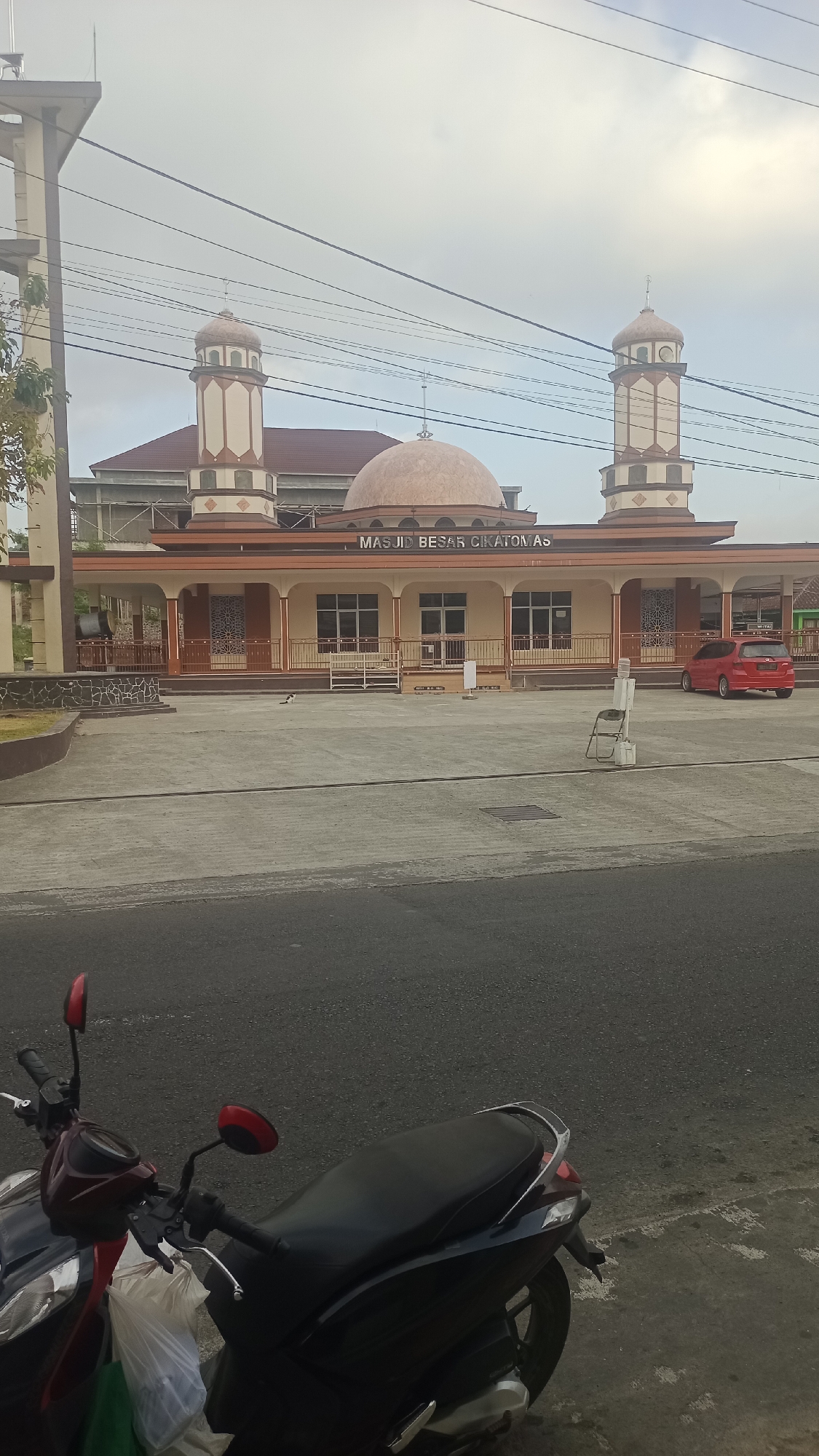 The image shows the exterior of the Grand Mosque of Cikatomas in Pakemitan, Indonesia, under a cloudy sky. The mosque features a large dome and two minarets, with the name of the mosque visible above the entrance. A red car is parked to the right of the mosque, and a motorcycle is parked in the foreground on the left. The mosque is set back from a paved road. The environment appears to be a public space with some vegetation visible to the left.