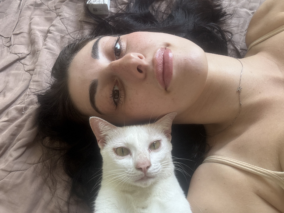 An overhead image depicts a light-skinned female individual and a white domestic cat lying on a textured light brown bedding or blanket. The scene is located in Cam Đức, Vietnam. The female, positioned on the right, has dark hair splayed around her head. Her face is directed towards the camera, with open brown eyes and slightly parted lips exhibiting a glossy texture. She wears a light-colored strap garment and a thin, silver-colored necklace with a small, star-shaped embellishment on her neck. The white cat, positioned to the left of the female, has light greenish-yellow eyes and a pink nose. Its head is tilted, facing the camera, resting adjacent to the female's left ear and shoulder area. Both subjects appear to be in a relaxed state.