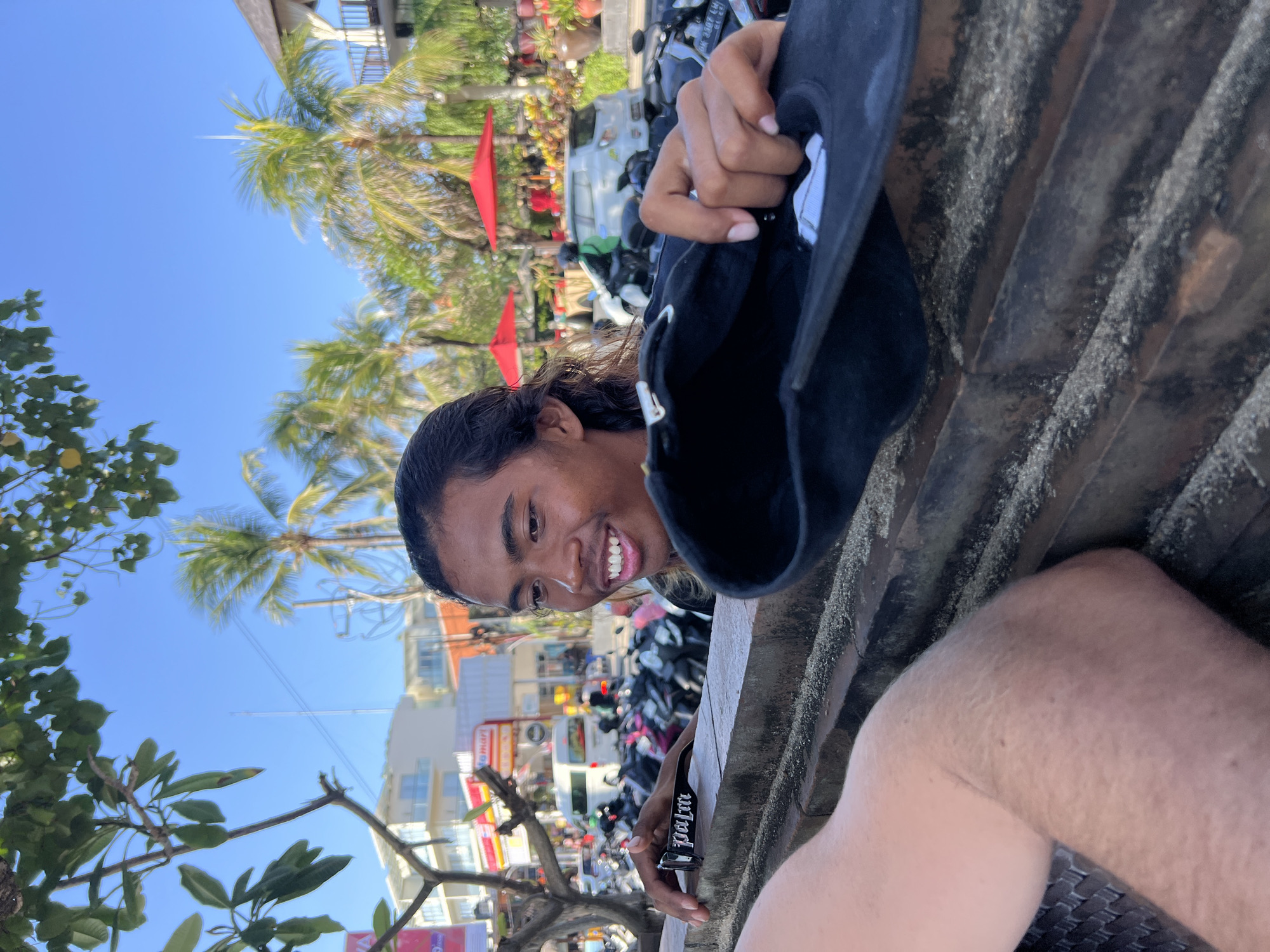 In Kuta, Indonesia, a young man with long dark hair and a bright smile leans over a stone wall, holding a black baseball cap. He is wearing a dark shirt, and a portion of another person's arm is visible in the foreground, resting on the wall. Behind him, a street scene unfolds with parked motorcycles, a white van, a building with balconies, and palm trees under a clear blue sky. Red umbrellas are visible in the background, suggesting an outdoor seating area. The overall impression is a candid moment captured in a bustling urban environment, likely a tourist area.