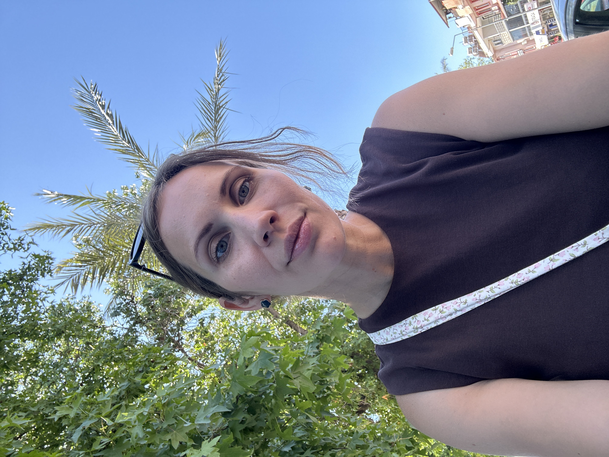 A low-angle selfie of a woman with fair skin, wearing a brown t-shirt with a strap across her chest. She has gray eyes and light brown hair pulled back. Sunglasses are perched on her head. Behind her, there are trees and a palm tree silhouetted against a clear blue sky. Buildings can be seen in the background. The location appears to be in an urban area of Fethiye, Turkey, based on the architectural style of the buildings and the presence of palm trees, which are common in that region.