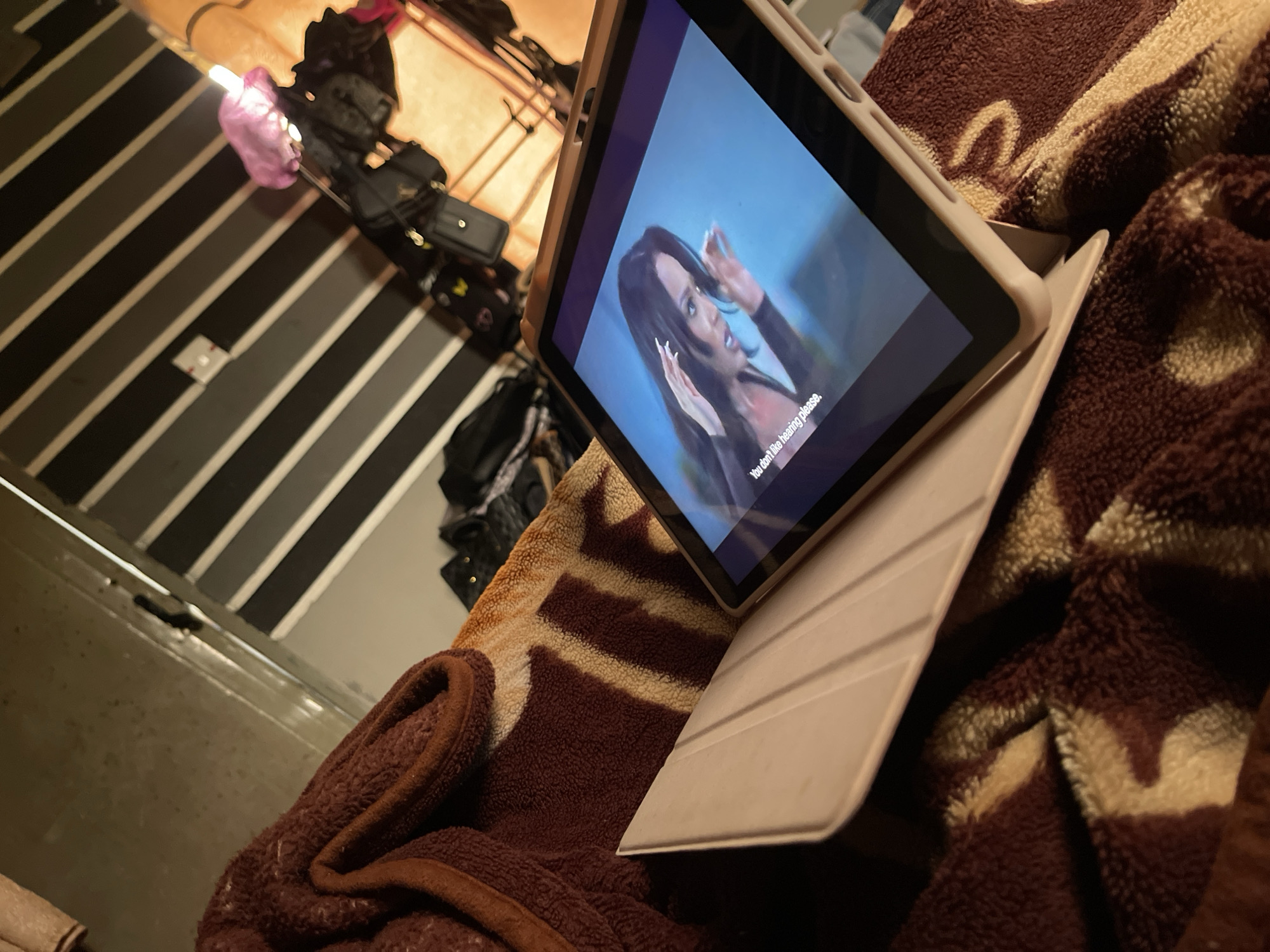 The image depicts a close-up, low-angle shot of a tablet computer resting on a patterned blanket. The tablet screen displays a woman with long dark hair and manicured nails, appearing to be in a scene from a video. White text, partially obscured, is visible at the bottom of the screen. The blanket has a textured, plush surface with a design featuring brown and beige patches. In the background, a wall with diagonal black and white stripes is visible, along with a light fixture and several dark-colored bags or clothing items hanging. The overall lighting suggests an indoor environment, possibly a bedroom or living area. The specific location in Rafin Par, Nigeria, cannot be determined from the visual information provided.