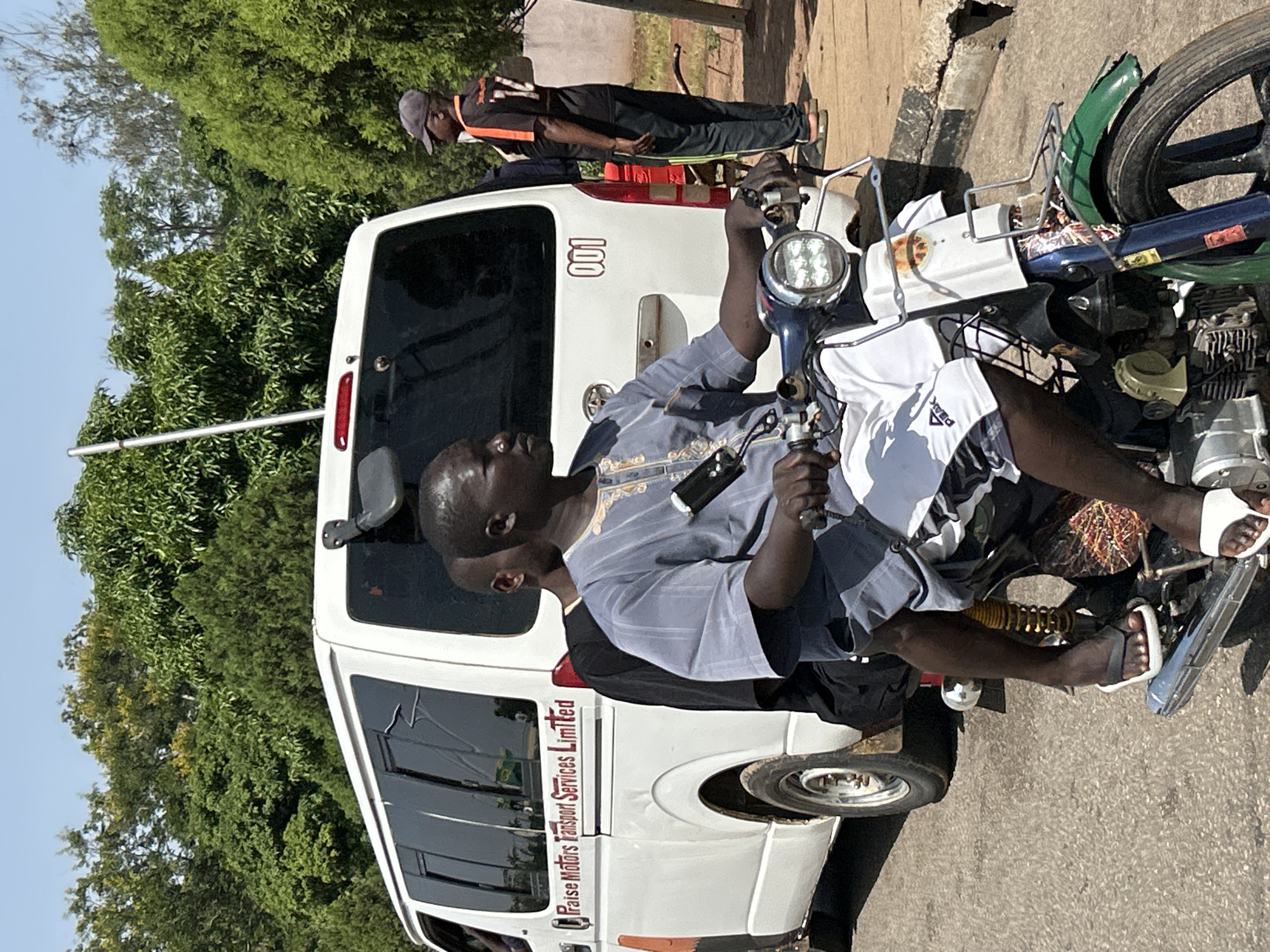 Two men are riding on a motorcycle, with the driver in the foreground and a passenger behind him. The driver is wearing a light blue, patterned tunic and white shorts. The passenger is wearing a black shirt and dark pants. They are traveling on a paved road, with a white van labeled "Praise Motors Transport Services Limited" to their left. The van has the number "001" printed on its rear. In the background, a man wearing a dark t-shirt with the number "17" on it and a baseball cap stands facing away from the camera. The environment appears to be a city street in Jos, Nigeria, with lush green trees lining the background and a clear, bright sky.