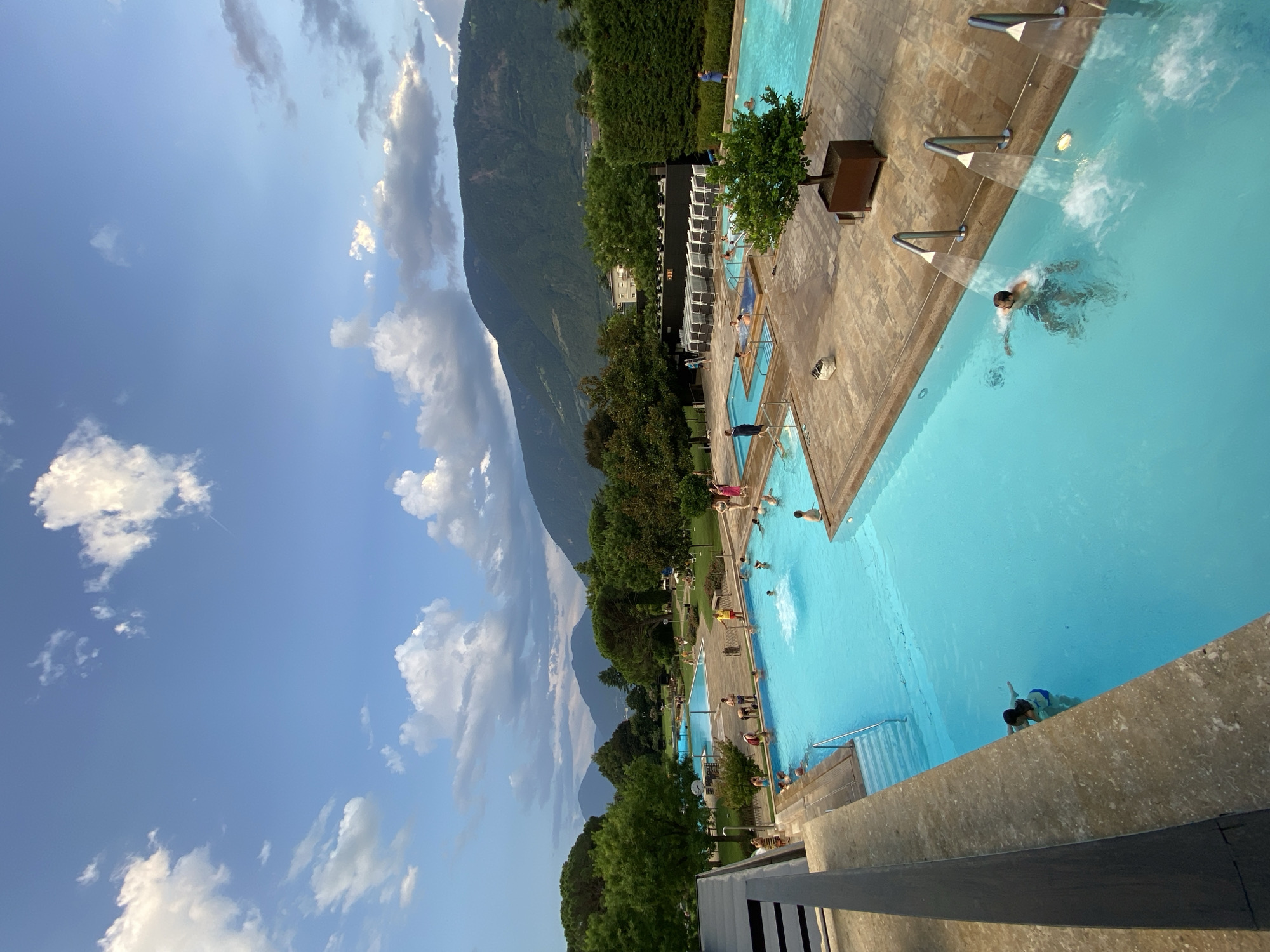 The image shows an outdoor swimming pool complex in Meran - Merano, Italy, under a bright, partly cloudy sky. The pools are filled with clear blue water and are surrounded by paved walkways and lounge areas. Several people are visible swimming, wading, and relaxing in and around the pools. Lush green trees and manicured hedges border the complex, with mountains in the background. The architecture of the facilities includes modern structures with glass elements and outdoor seating. Water jets are active in one section of the pool, creating a dynamic visual effect. The scene suggests a recreational and leisure setting, possibly a public park or a spa.