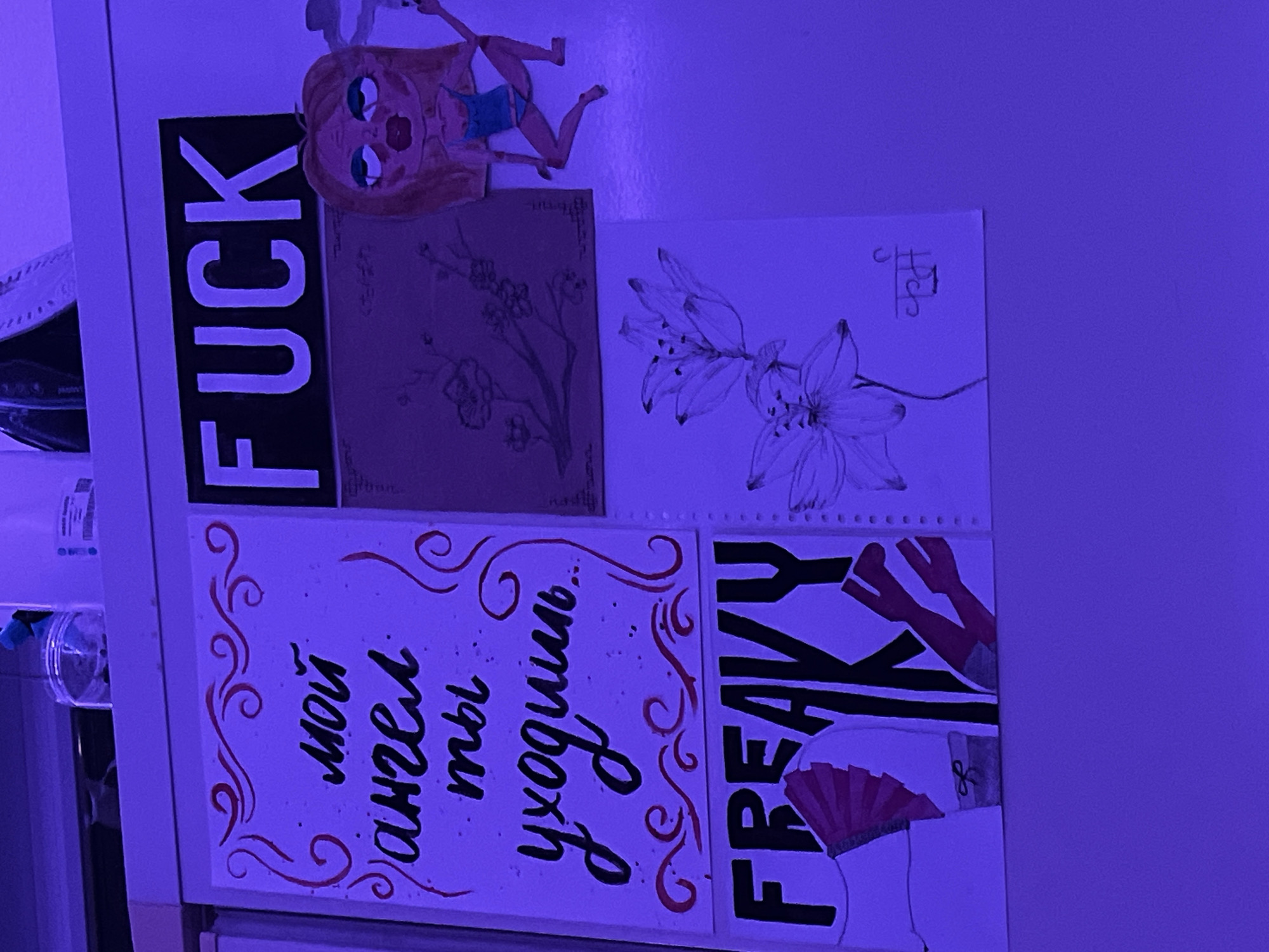 The image shows a refrigerator door covered with various drawings, paintings, and a small doll. From left to right, top to bottom, the items include: a black and white sign with the word "FUCK" in large block letters; a stylized drawing of a doll-like figure in a blue swimsuit; a sepia-toned drawing of blossoming branches; a black and white drawing of a lily; a white card with the Russian phrase "мой ангел ты уходишь..." (my angel, you are leaving...) in stylized lettering; and a drawing of a woman's lower body with a fan and the word "FREAKY" in block letters. The image is illuminated with a strong, possibly artificial, purple light. There are no immediate indicators of the location being Saint Petersburg, Russia, nor are there any people visible in the image.