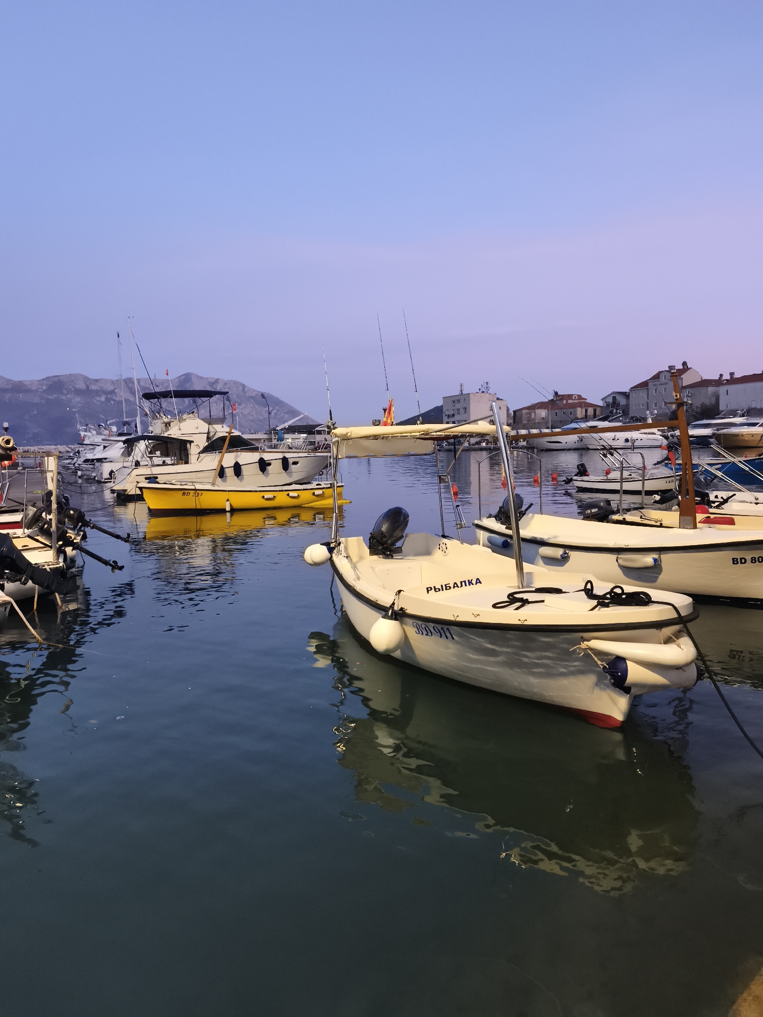 The image shows a harbor in Budva, Montenegro at dusk. Several boats are docked, including a yellow one and several white ones. The boat closest to the camera has the word "РЫБАЛКА" written on its side. Buildings are visible in the background, as are mountains in the distance. The water is calm, reflecting the colors of the sky and the surrounding objects.