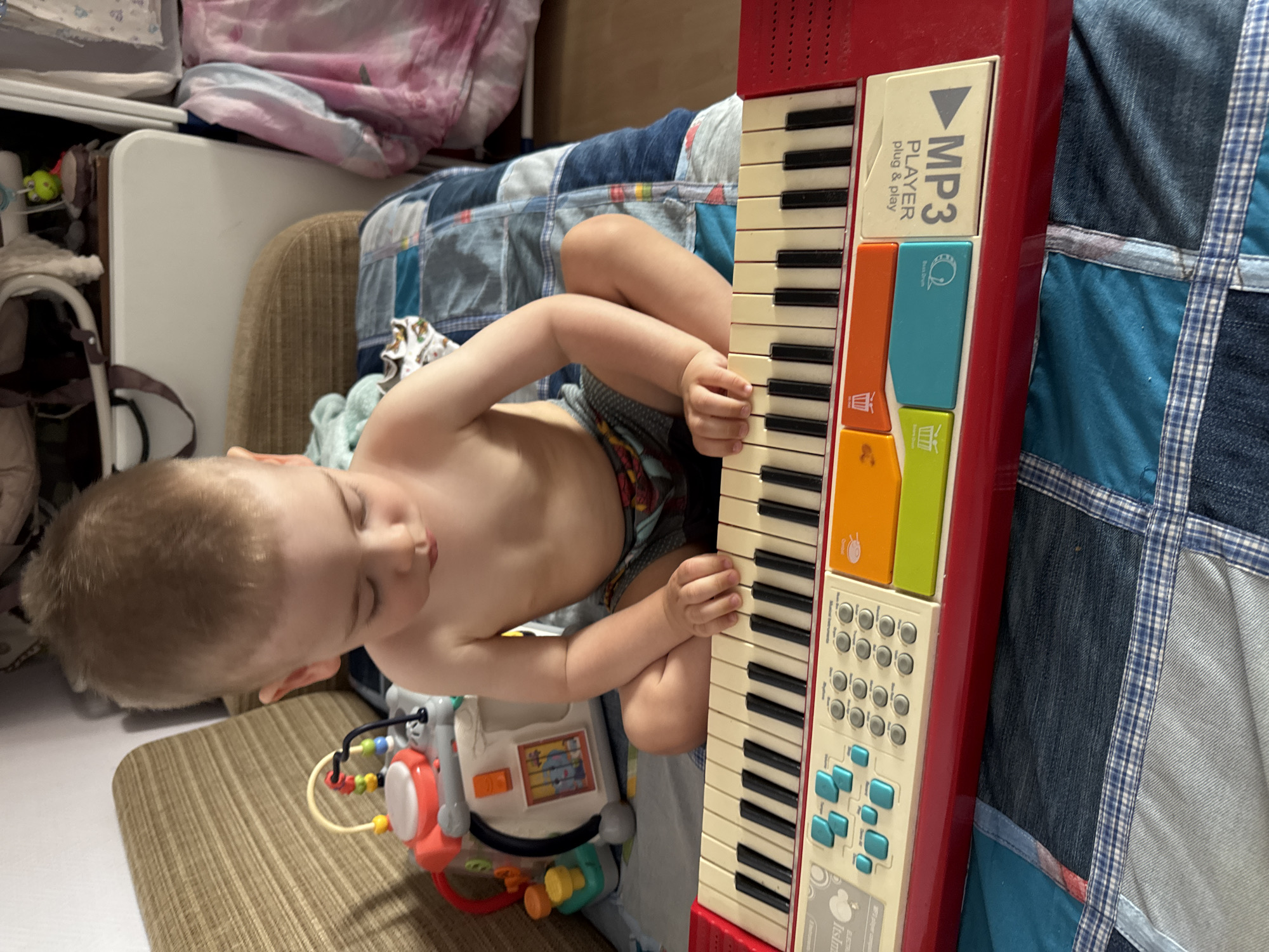 The media file shows a young child, likely a toddler, lying on their stomach and playing a red toy keyboard. The child is looking down at the keyboard, with their right hand resting on the keys. The keyboard has black and white keys, with colorful buttons and a white panel that says "MP3 PLAYER plug & play". The child is wearing patterned shorts. The scene appears to be indoors, with a bed or sofa covered in a patchwork quilt in the background. There are also some other toys visible near the child. The lighting suggests it is daytime. The overall mood seems relaxed and playful. There is no indication of the time of day, weather, or specific location within Ufa, Russia, beyond the general context provided.