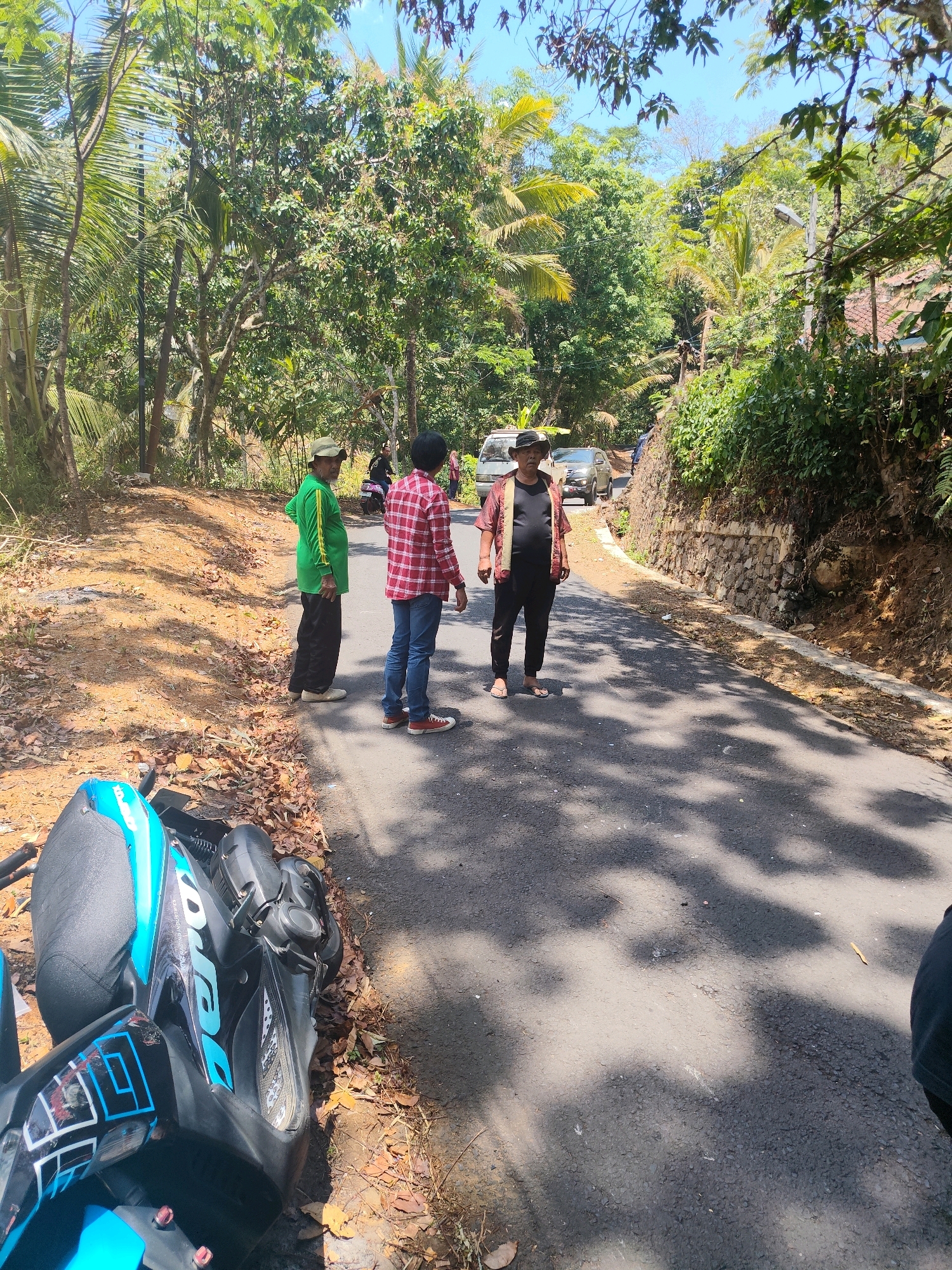 The image shows a paved road winding through a rural area with dense vegetation and trees on either side, under a clear blue sky. In the foreground, a blue and black scooter is parked on the left side of the road, partially on a dirt and leaf-strewn shoulder. On the road, three individuals are visible. A person wearing a green long-sleeved shirt and black pants stands facing away from the camera, looking towards the right. To their right, a person in a red and white checkered long-sleeved shirt and blue jeans stands facing the camera. Further down the road to the right, another person wearing a dark shirt, a patterned shawl or sash draped over their shoulders, dark pants, and a hat is standing and looking towards the camera. In the background, more trees and foliage are present, and two vehicles, a white van and a silver car, are parked on the road. The location is identified as Karangjaya, Indonesia. The scene appears to be a roadside gathering or stop in a tropical, semi-rural environment.