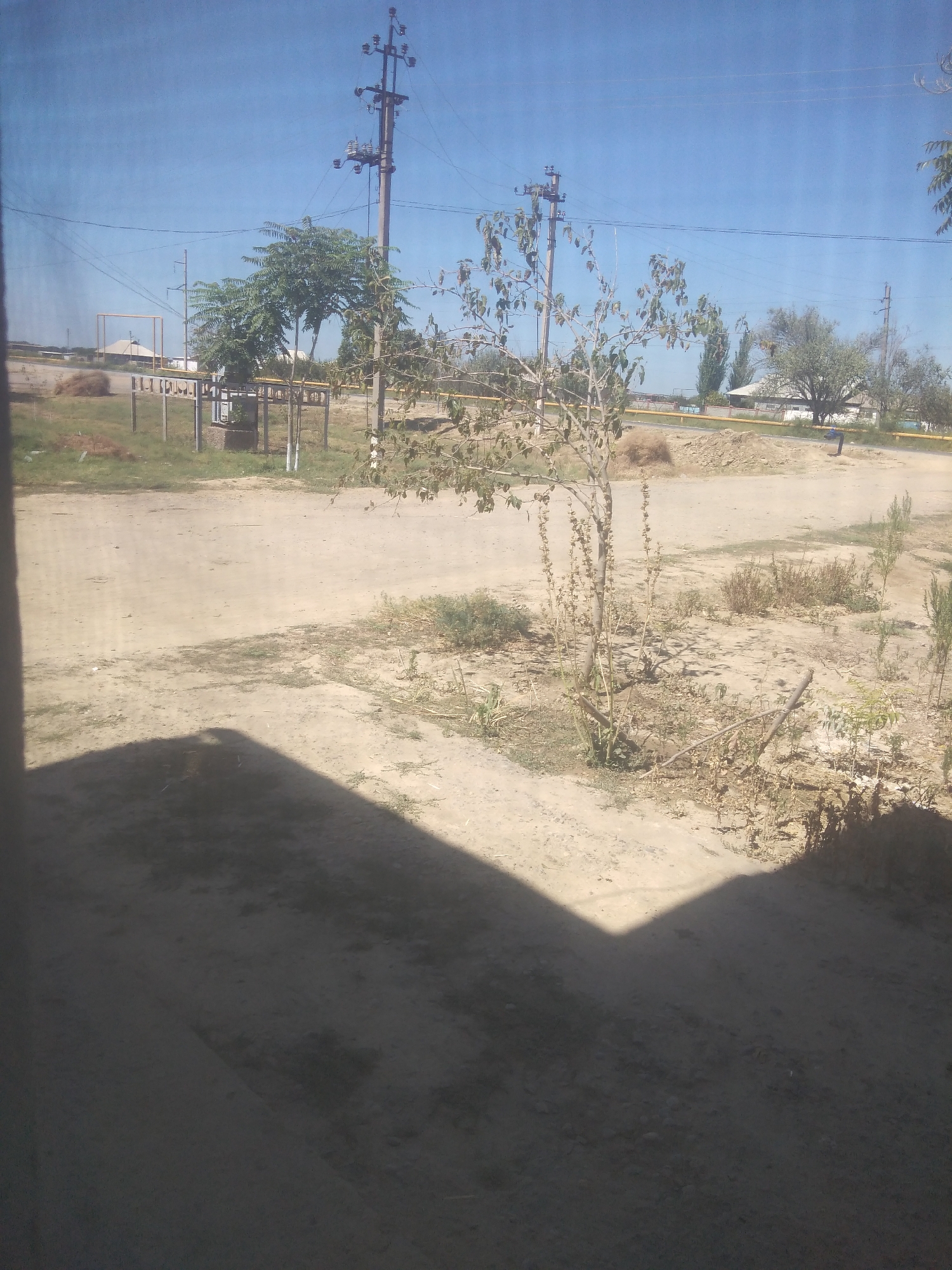 An outdoor, eye-level shot captures a dusty, unpaved area with sparse vegetation under a clear blue sky. In the background, a tall utility pole with electrical equipment stands prominently. To the left of the utility pole, a metal structure with yellow beams is visible. Further in the distance, several trees with lush green foliage are scattered across the landscape. The ground is a light brown, dusty soil, with patches of dry grass and weeds. A large, dark shadow, possibly from a building or object not fully in frame, covers a significant portion of the foreground, obscuring details on the ground. A thin, bare-branched tree stands in the middle ground, casting a dappled shadow on the ground. The overall impression is one of a rural or semi-developed area.