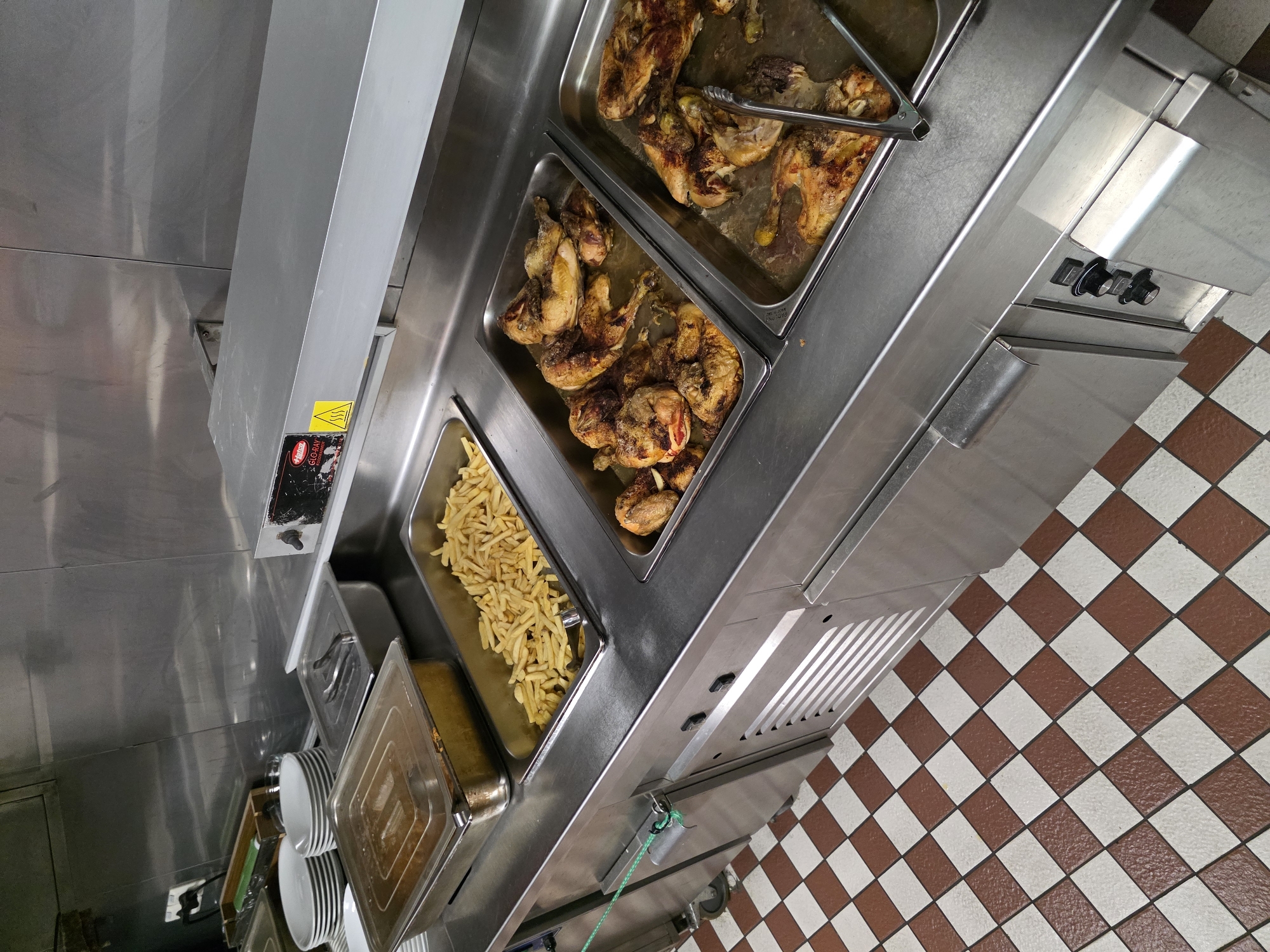 This is a forensic, matter-of-fact description of a commercial kitchen serving area in Brindisi, Italy. The scene depicts a stainless steel buffet counter filled with food. On the left, a stainless steel container holds what appears to be a stew or saucy dish, covered with a clear lid. Adjacent to it, a deep stainless steel tray is filled with a generous portion of golden-brown french fries, with a pair of stainless steel tongs resting inside. To the right of the fries, two more deep stainless steel trays contain roasted chicken pieces. The chicken is golden brown and appears seasoned, with some pieces showing char marks from roasting. The tray on the left holds more chicken pieces than the tray on the right. The counter is clean and reflects the overhead lighting. In the background, a stainless steel wall paneling and a food warming lamp with warning labels are visible. The floor is tiled with a red and white checkered pattern. No people are present in the immediate scene.