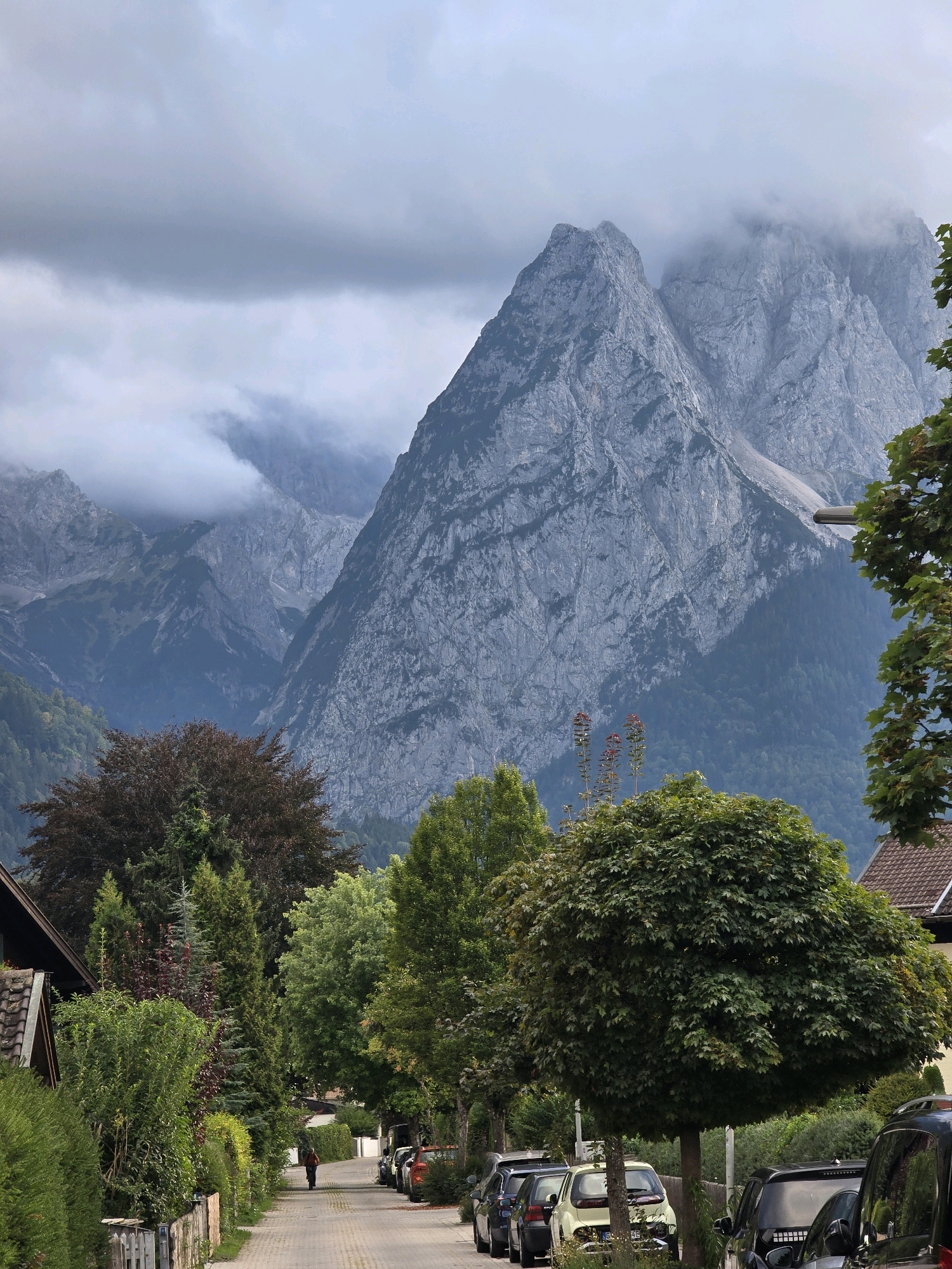 The image depicts a street in Garmisch-Partenkirchen, Germany, with a person on a bicycle in the distance. The street is paved with cobblestones and lined with trees and hedges on both sides. Several parked cars are visible along the right side of the street. In the background, imposing, jagged mountains rise under a cloudy sky, their peaks partially obscured by mist. The overall scene suggests a somewhat overcast day in a picturesque, mountainous town.