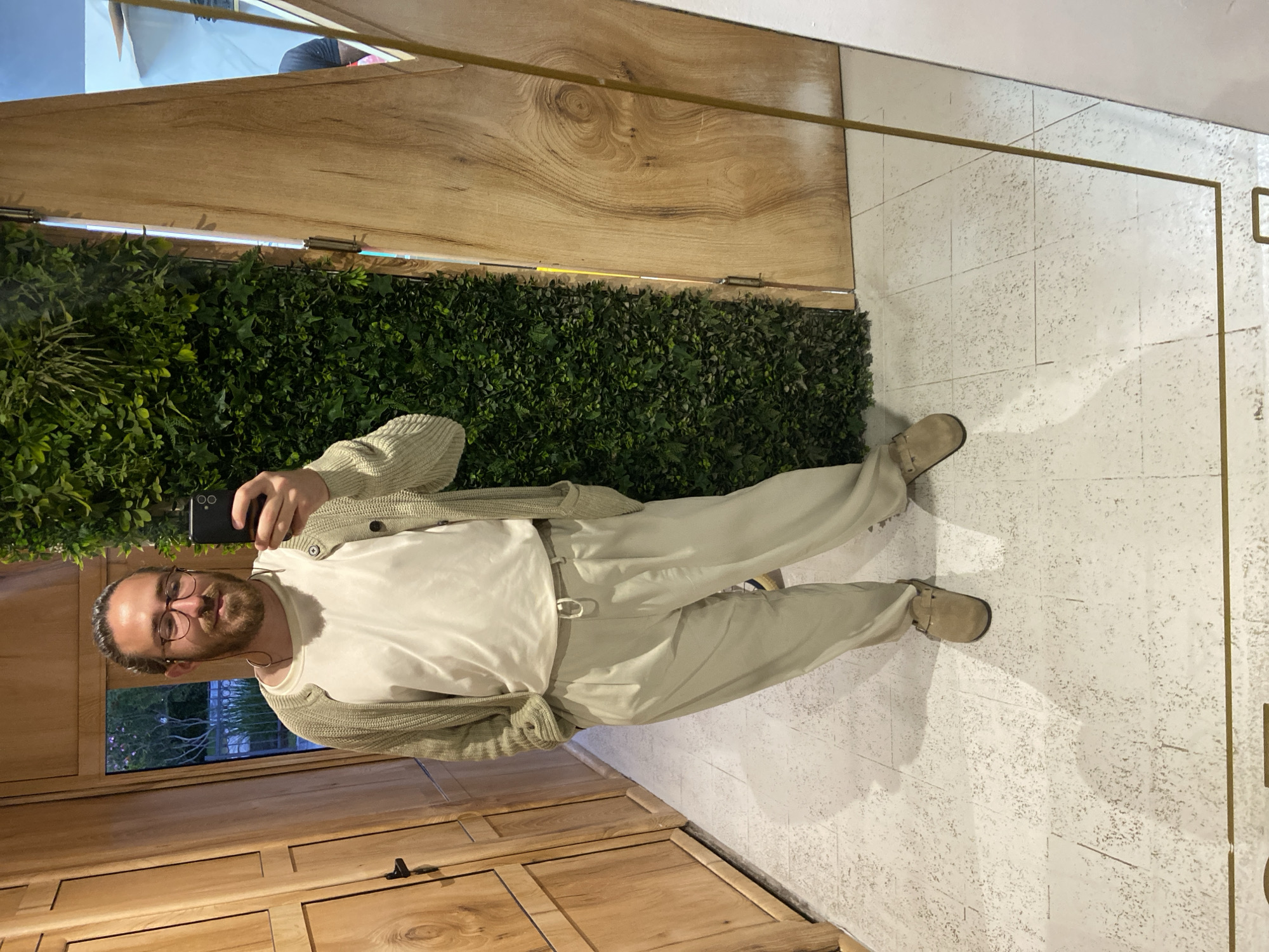 A man with a beard and glasses is taking a selfie in a mirror. He is wearing a white t-shirt, khaki pants, a light cardigan, and tan shoes. The reflection shows the man standing in front of a wooden wall, a vertical garden, and a wooden cabinet. The mirror is framed with a gold metal trim. The background appears to show Petrovac centar, Montenegro.
