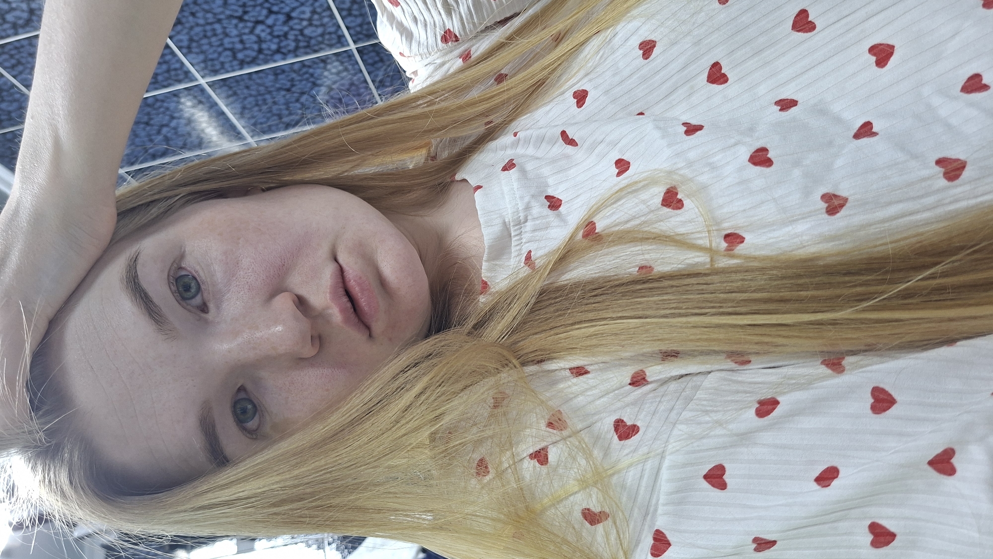 A young, fair-skinned woman with blonde hair is lying down, her head slightly tilted, with her right hand raised near her forehead. She is wearing a white blouse with small red heart patterns. The background includes a grid-like tile pattern, suggesting an indoor location near a window. It is unclear if the location is in the city of Chaikovsky, Russia.