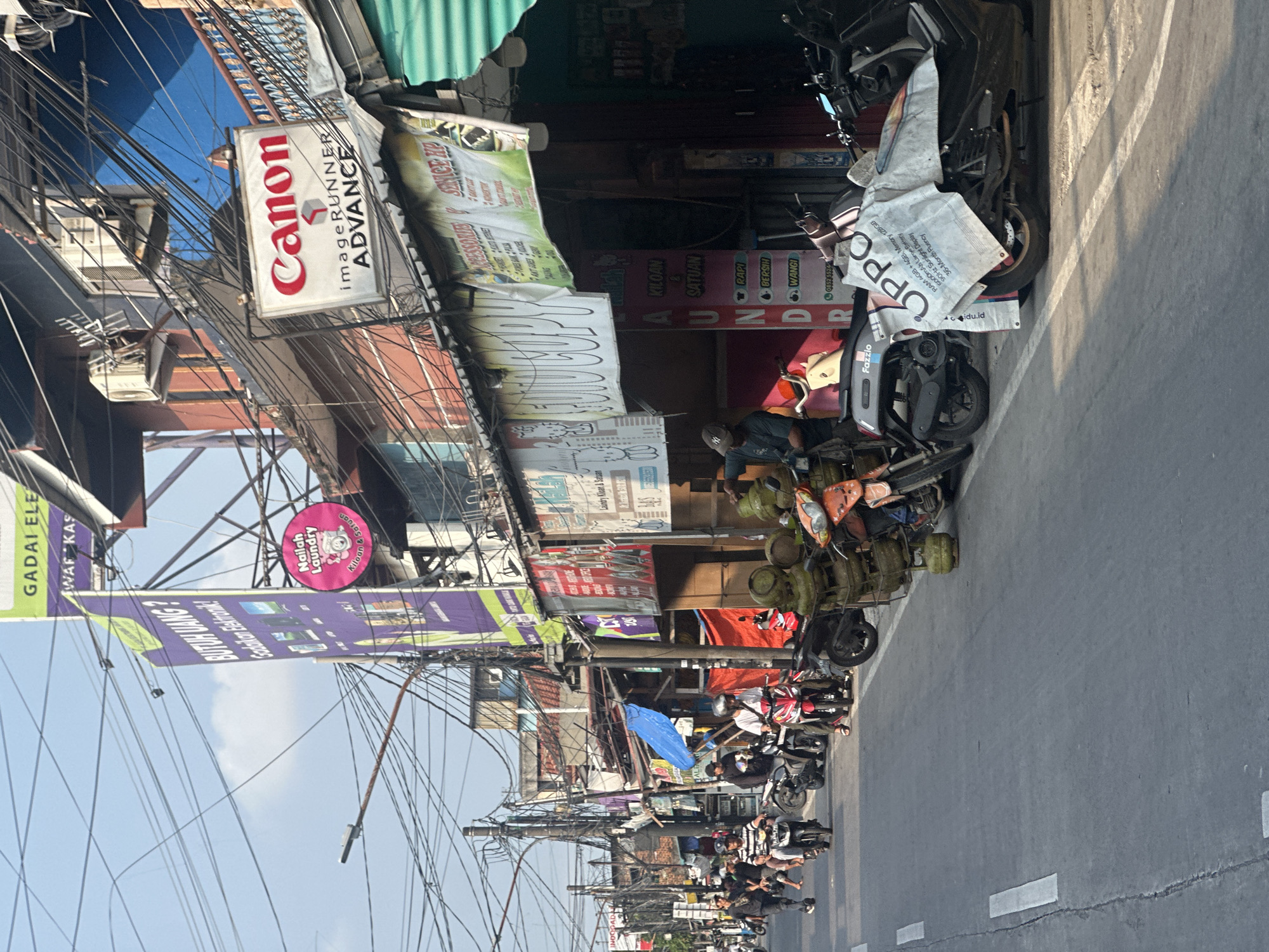 The image captures a street scene in North Jakarta, Indonesia. The view is from a street-level perspective, focusing on a line of parked motorcycles along the right side of the road. Above the motorcycles are shopfronts crowded with signage. Notably, there's a "Canon imageRUNNER ADVANCE" sign, various laundry advertisements, and a banner advertising "GADAI ELE" (pawn shop) and electronic goods. Electrical wires are tangled overhead, obscuring parts of the sky. Several people are visible near the motorcycles. The overall impression is a typical urban street scene with a focus on business activity and transportation.
