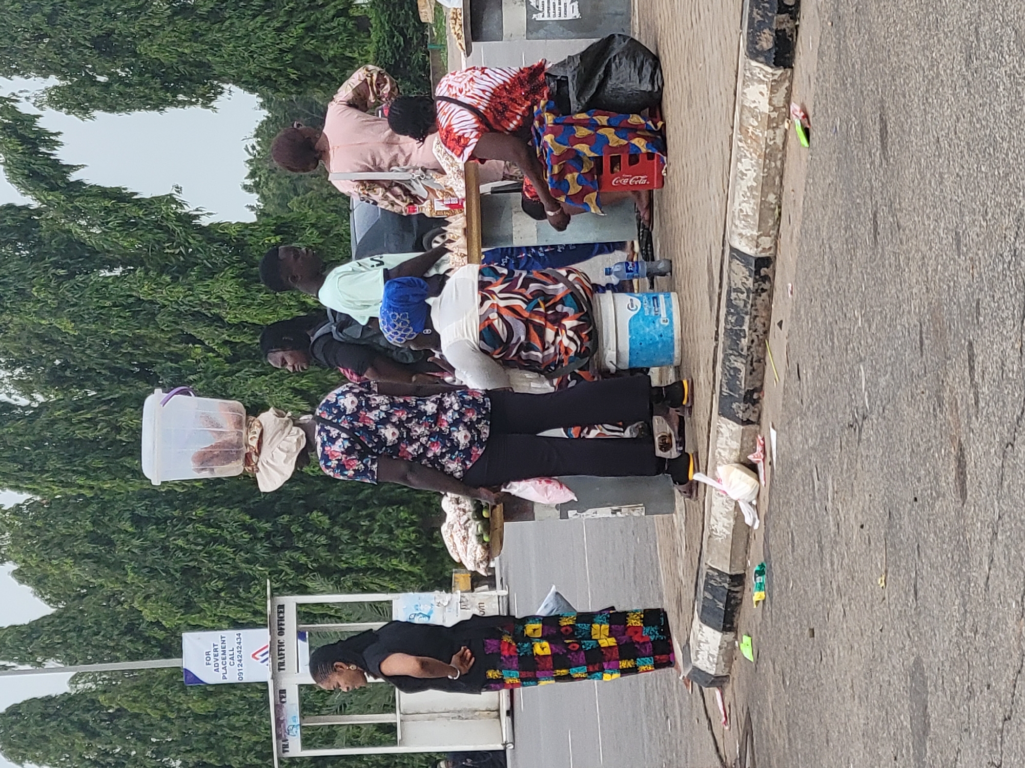 The image depicts a bustling street scene in what appears to be Abuja, Nigeria, featuring multiple individuals engaged in street vending activities. The setting is a paved road alongside a concrete sidewalk, bordered by lush green trees in the background. The sky is bright, indicating daytime.
In the foreground on the sidewalk, a woman with dark hair wears a black top and a colorful, multi-patterned long skirt. Further back, another woman is seen carrying a large, clear plastic container on her head, suggesting she is a hawker or vendor. She wears a floral shirt and dark trousers.
To the right, a group of people, including men and women, are gathered around what appear to be makeshift stalls or selling points on the sidewalk. One woman, wearing a red and white patterned outfit, is seated next to a red Coca-Cola crate, which contains bottles. Other individuals are also seated or standing, with various items like water bottles, buckets, and bags visible around them, implying they are also vendors or customers. A man in a light green shirt is visible among this group.
The concrete sidewalk has a curb painted with faded black and white stripes, separating it from the gray asphalt road. There is a noticeable amount of litter, including plastic bags and wrappers, scattered on both the sidewalk and the street.
A white sign, partially visible on the left side of the image, displays text that reads "FOR ADVERT PLACEMENT CALL 09124242434" and also "TRAFFIC OFFICER" at its bottom. The scene captures a typical daily commerce environment in an urban or semi-urban area.