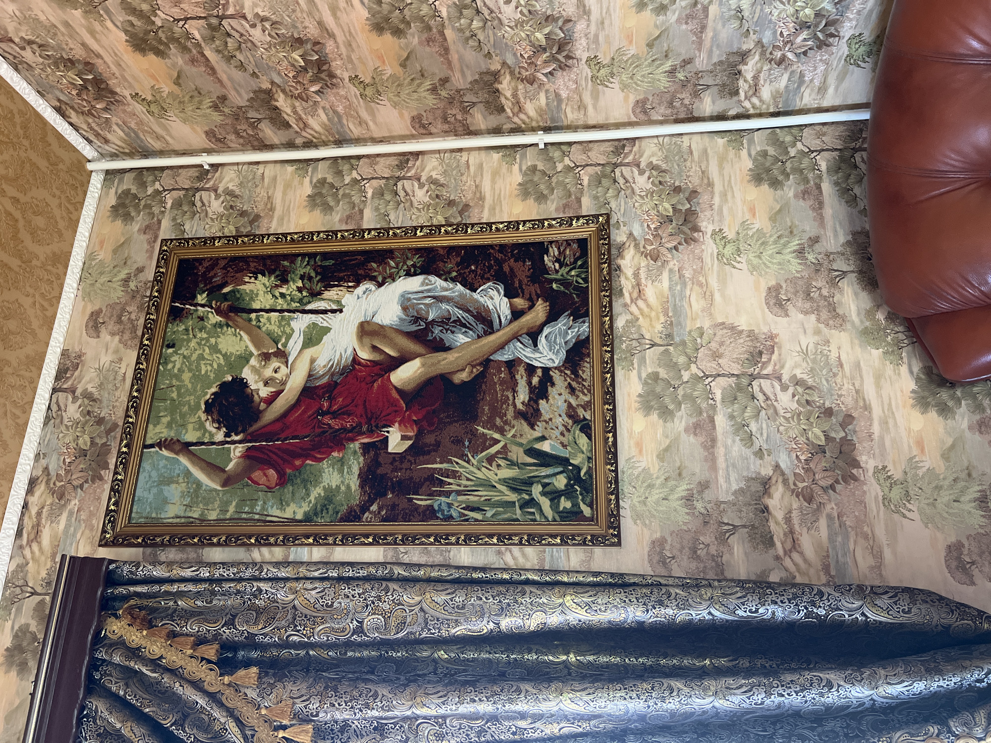 This media file is a photograph taken in Krasnoe-na-Volge, Russia. The image captures a corner of a room, focusing on a tapestry hanging on a wall and the ceiling above it. The tapestry, framed in an ornate gold-colored border, depicts a scene with two figures. A male figure, dressed in red, is embracing a female figure, who is draped in white. They are positioned in a naturalistic setting, with hints of greenery and possibly a body of water. The figures appear intertwined, and their poses suggest an intimate or dramatic moment. The tapestry's style is reminiscent of classical or romantic art.
The wallpaper on the wall and ceiling features a repeating pattern of trees and rocky landscapes, rendered in muted, earthy tones of green, brown, and beige. This wallpaper creates a continuous, immersive natural scene that extends to the ceiling, which is also covered in the same patterned material. A white trim separates the wall and ceiling, and a thin white pipe or conduit runs along this junction.
In the foreground, a dark, richly patterned fabric is visible, likely curtains or upholstery, with intricate paisley designs in black, gold, and silver. Tassels and decorative trim in gold are attached to this fabric, adding a touch of opulence. To the right of the frame, a portion of a brown leather sofa is visible, indicating a furnished living space. The lighting suggests it is daytime, possibly indoors with natural light. There is no visible text in the image.