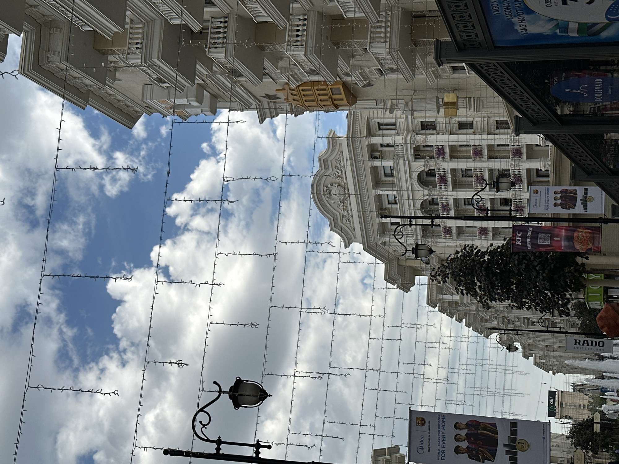 The image shows a street scene in Tashkent, Uzbekistan, possibly on Amir Timur Square or a nearby avenue, characterized by ornate architecture and festive decorations. On the right, a modern building with multiple balconies and detailed white stone accents dominates the frame. The building is adorned with strings of small lights, suggesting a festive or decorative purpose. To the left, another building with a classical facade featuring columns and arched windows is visible.
Street furniture includes decorative lampposts and hanging strings of lights that stretch across the street, creating a canopy effect. Several banners hang from the lampposts and buildings. One prominent banner on the left displays the "Midea" logo and advertises "FC Barcelona," featuring images of three football players. Another banner advertises "KFC Shefburger." A dark green tree is positioned in the mid-ground.
In the lower part of the image, elements of urban infrastructure are visible, including a fountain with water jets, and more banners, one of which reads "RADO SWITZERLAND." A person wearing an orange hat is partially visible in the foreground, to the left of the RADO banner. A large blue and white advertisement board is on the right side, featuring text in Uzbek and imagery related to Uzbekistan's national symbols, such as the flag. The sky is partly cloudy with a mix of blue sky and white clouds. The overall impression is of a well-maintained urban environment, possibly a shopping or entertainment district, decorated for an occasion.