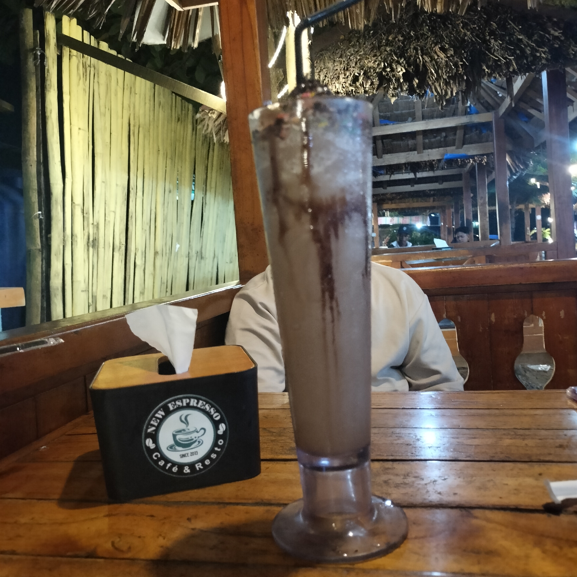 The scene is at New Espresso Café & Resto in Titibaro, Indonesia, as indicated by the tissue box on the wooden table. The table also holds a tall glass of chocolate beverage with a straw. Behind the glass, a person is seated at the table. The structure appears to be an open-air pavilion with a thatched roof. The background features bamboo fencing. Other people are visible in the distance through wooden beams. The lighting suggests it is nighttime.