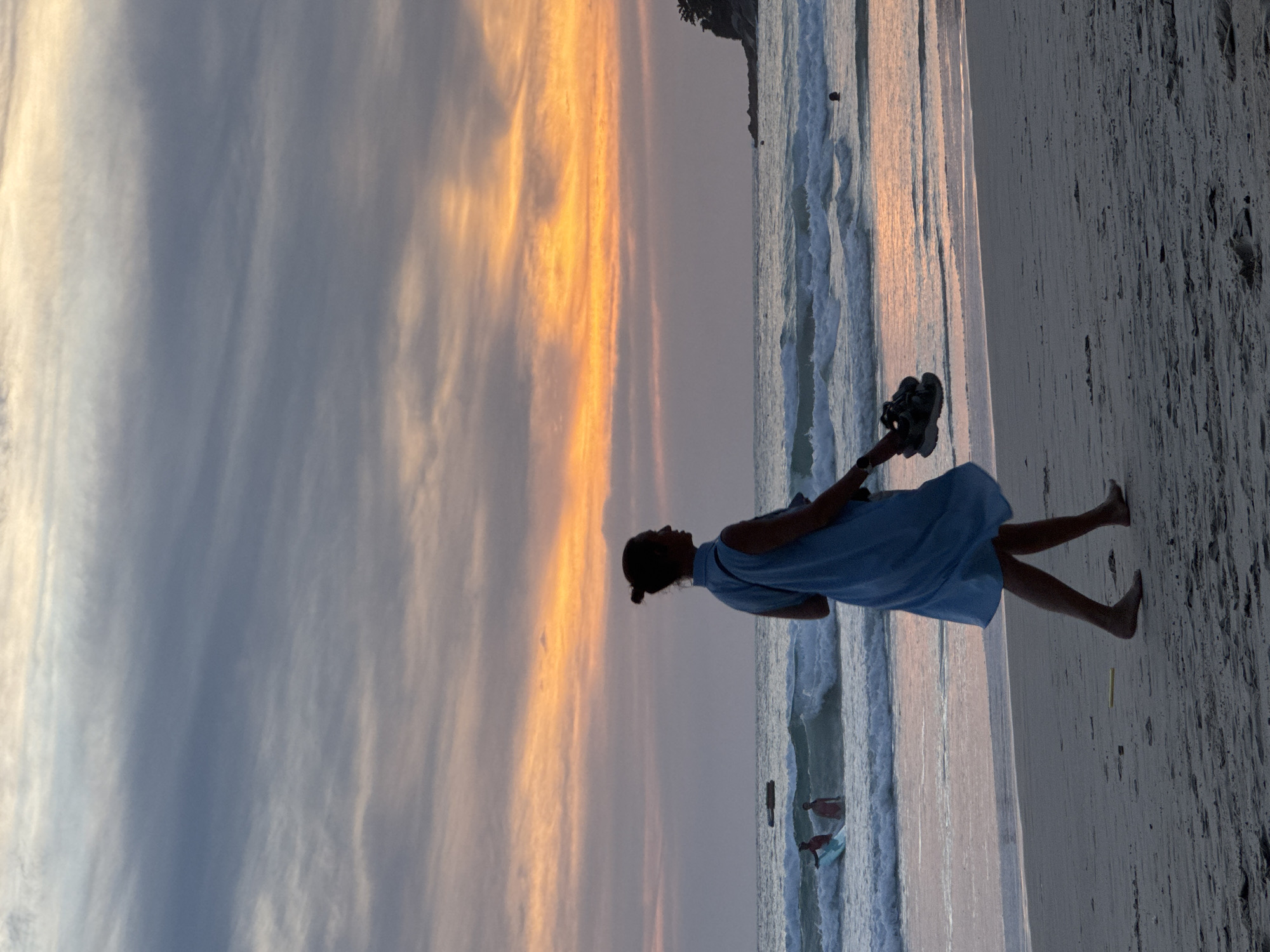 A silhouette of a woman in a light blue dress walks along the sandy shore of Karon Beach, Thailand, at dusk. She holds a pair of dark sandals in her right hand. The ocean stretches out before her, with waves gently rolling onto the beach. In the distance, a few individuals are visible in the water, one with a surfboard. The sky above is a canvas of dramatic clouds, transitioning from soft blues and grays to vibrant oranges and yellows as the sun sets. A rocky outcrop with lush vegetation juts out into the sea on the right side of the frame. The overall scene captures a serene moment at the end of the day.