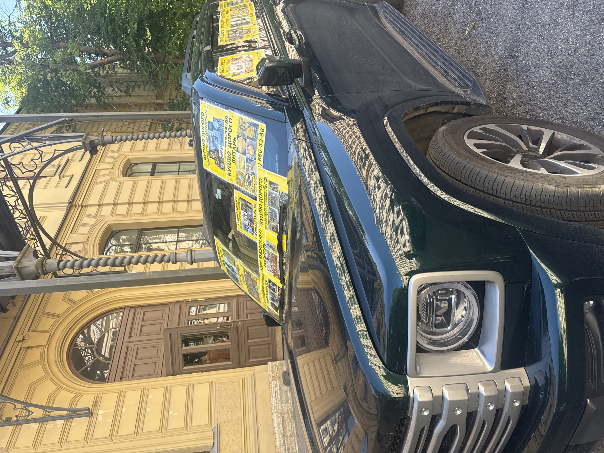 This media file is an image of a dark green SUV parked on a street. The vehicle is covered in bright yellow flyers that advertise a service for buying valuable items. The flyers, written in Russian, feature text such as "КУПЛЮ ДОРОГО" (I will buy for a high price), listing items like amber, porcelain figurines, glassware, sculptures, and jewelry from the USSR. Phone numbers are prominently displayed on the flyers, indicating contact information for the service.
The car is parked in front of a building with a yellow facade, featuring large arched windows and ornate architectural details. The building's reflection is visible on the polished surface of the car. A tree with lush green foliage extends over the scene from the left, suggesting it is daytime and likely spring or summer. The overall lighting indicates it's a sunny day. The ground appears to be asphalt. The image is framed at an angle, focusing on the front and side of the car and the building it's parked next to. The scene is set in Saint Petersburg, Russia.