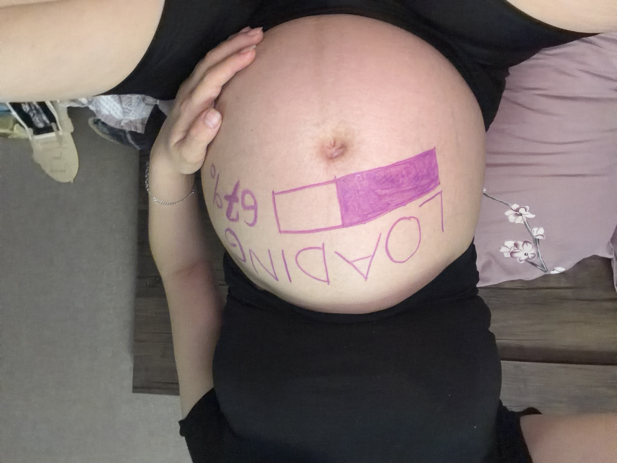 A pregnant person is lying on a bed, wearing a black t-shirt and black shorts. Their pregnant belly is decorated with purple marker. The writing on the belly says "LOADING" above a progress bar that is about two-thirds filled. To the right of the progress bar, "67%" is written. The person is holding their belly with their right hand, and a silver bracelet is visible on their left wrist. The background includes a wooden headboard, a pink pillow with a floral pattern, and a grey wall. There is also an object that appears to be a shoe or slipper on the right side of the frame. The scene is indoors, likely a bedroom.