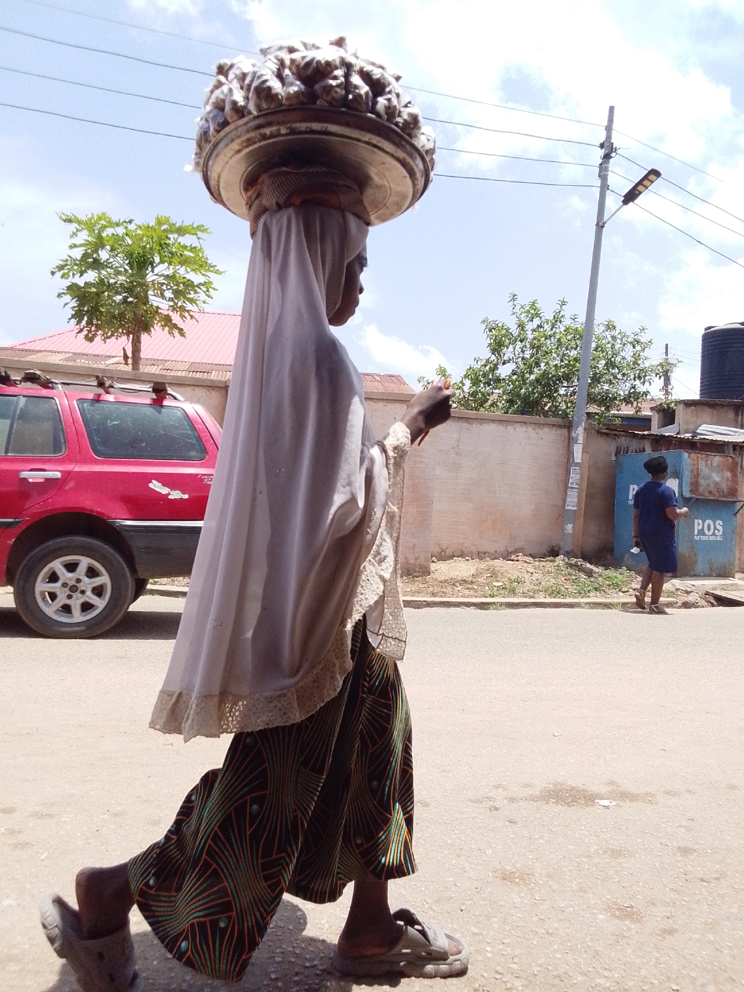 A woman walks on a paved road in Jos, Nigeria. She carries a large, round tray filled with what appears to be food items on her head. She is wearing a light-colored headscarf and a long, patterned skirt. Another woman is visible in the background walking toward a "POS" (Point of Sale) kiosk. A red SUV is parked on the side of the road. The sky is partly cloudy. A power line runs above the street.