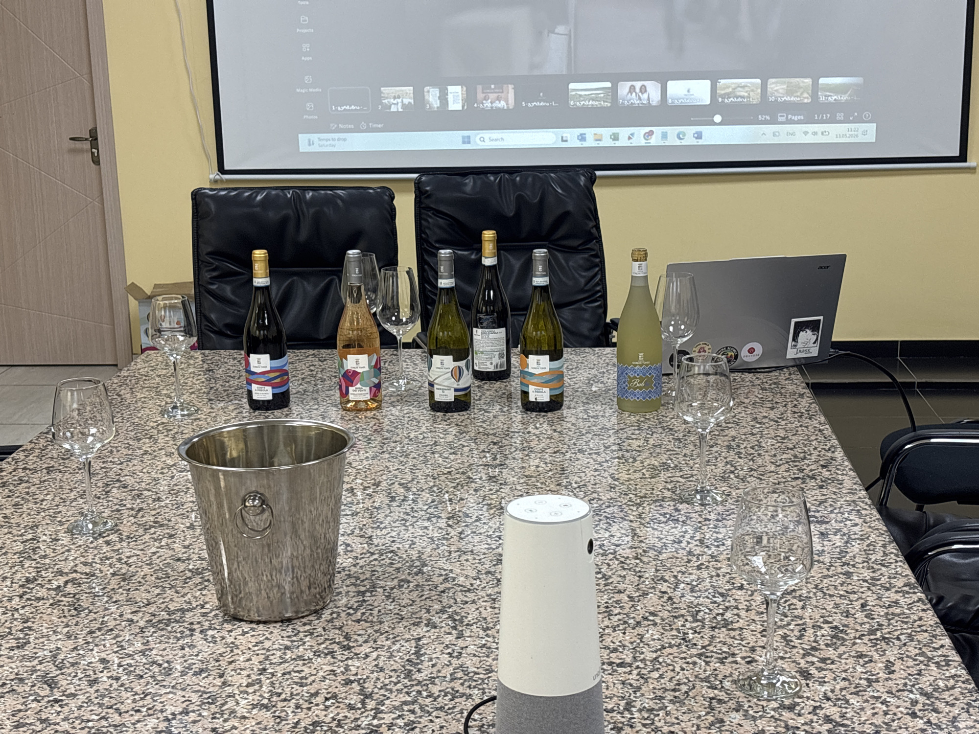 The image shows a meeting or presentation setup on a patterned table in a room. Several wine bottles and wine glasses are arranged across the table. Behind them are two black office chairs and a projector screen displaying a computer interface. A silver ice bucket is placed near the center of the table, and a white smart speaker sits in the foreground. An Acer laptop is visible near the right side of the table. The overall lighting suggests an indoor setting, likely in an office or conference room. There is no indication of the location in Tbilisi, Georgia.