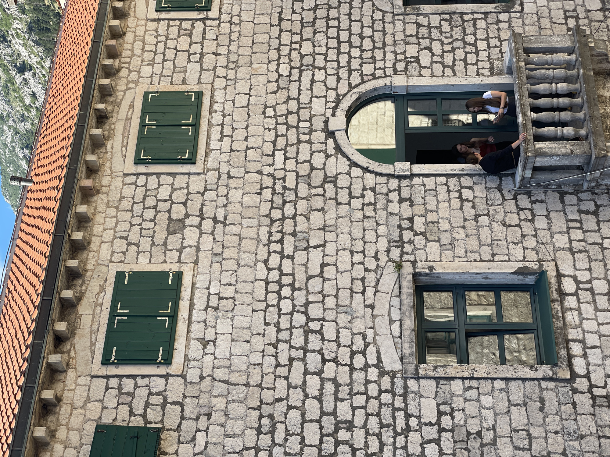 The image shows a stone building with a red tiled roof, located in Kotor, Montenegro. The building has several windows, some with green shutters and others with green frames. On a balcony on the second floor, three young adults are visible. One individual, wearing a black shirt, stands to the left, facing two others. A woman in a red top stands to the right of the man in black, and a woman in a white top stands further to the right, looking at her phone. The balcony is made of stone and has a railing with balusters. The background shows a mountainous landscape, characteristic of Kotor.