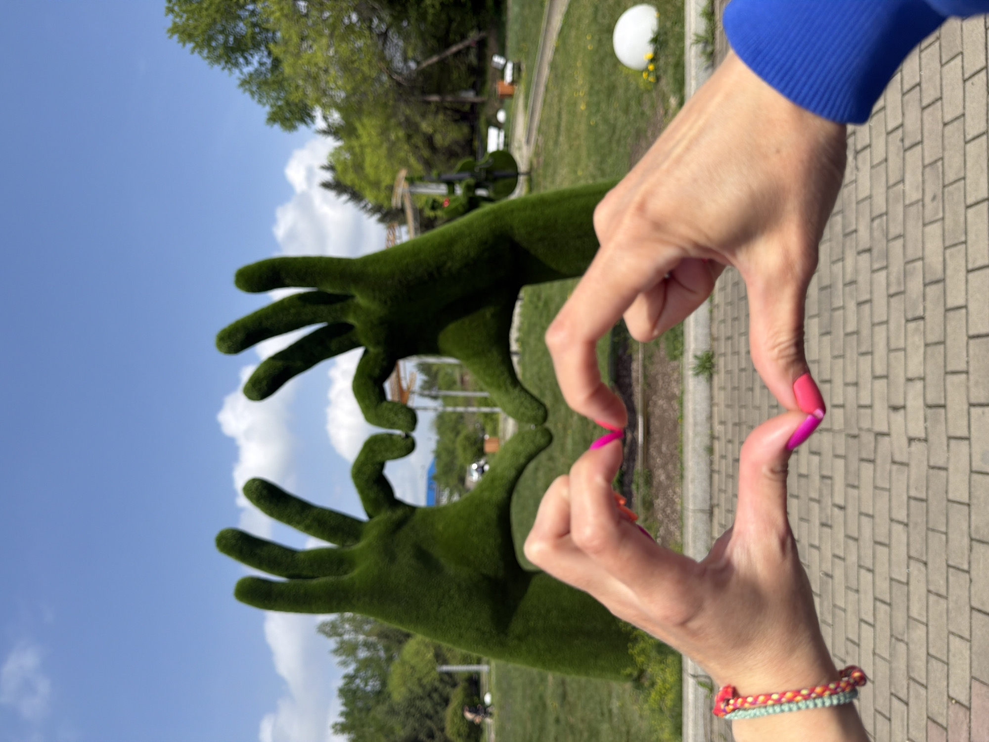 The image shows a view in what appears to be a park or garden setting, likely in Birobidzhan, Russia. Two large topiary hands, covered in green grass-like material, are positioned opposite a person making a heart shape with their hands. The person has fair skin and pink nail polish. The hand on the right is wearing a multi-colored bracelet. The background features trees, lawn, a light-colored pavement with brick-like patterns, and what appears to be other topiary sculptures. The sky is blue with some clouds.